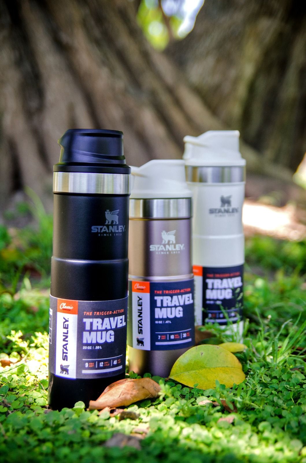Tres tazas de viaje Stanley en negro, tostado y blanco apiladas sobre el césped frente a un árbol.