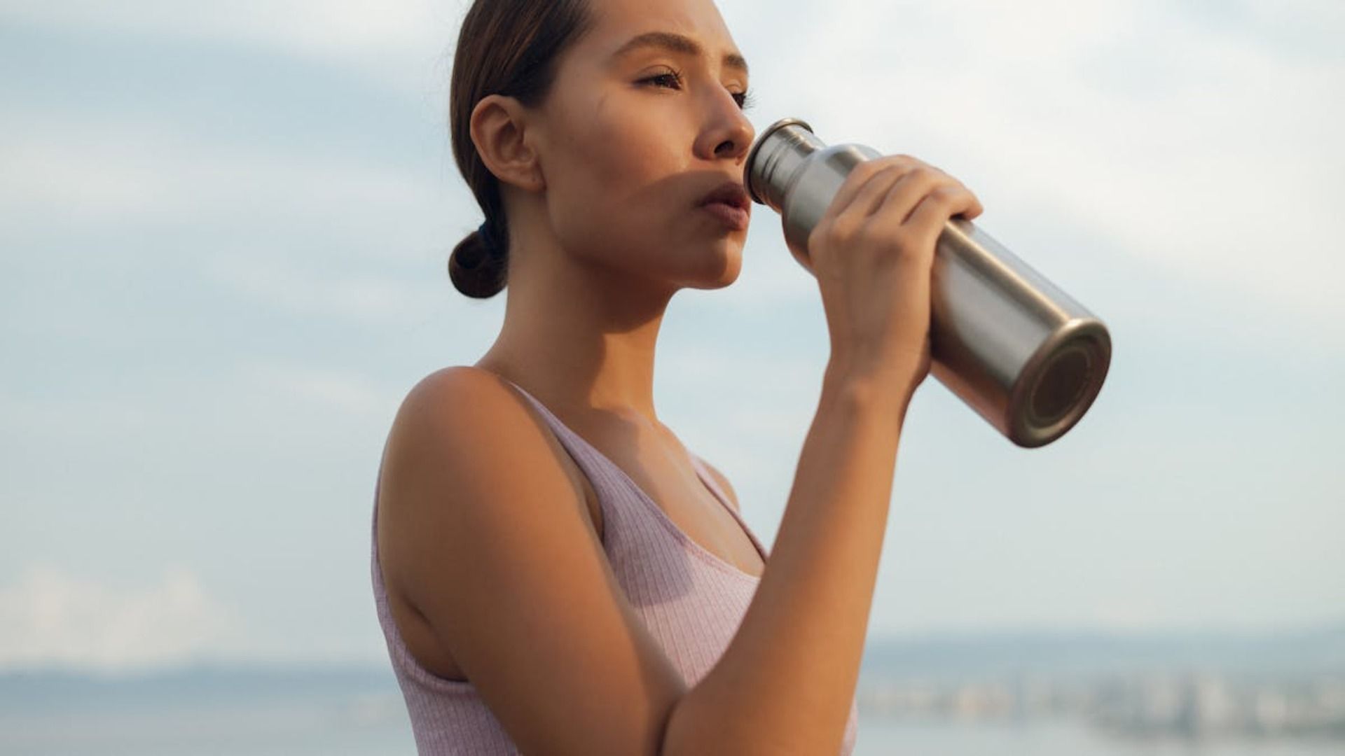 Una mujer bebe de una botella de agua plateada al aire libre, bajo la luz del sol. Lleva una camiseta rosa claro sin mangas y mira a un lado con la boca abierta.