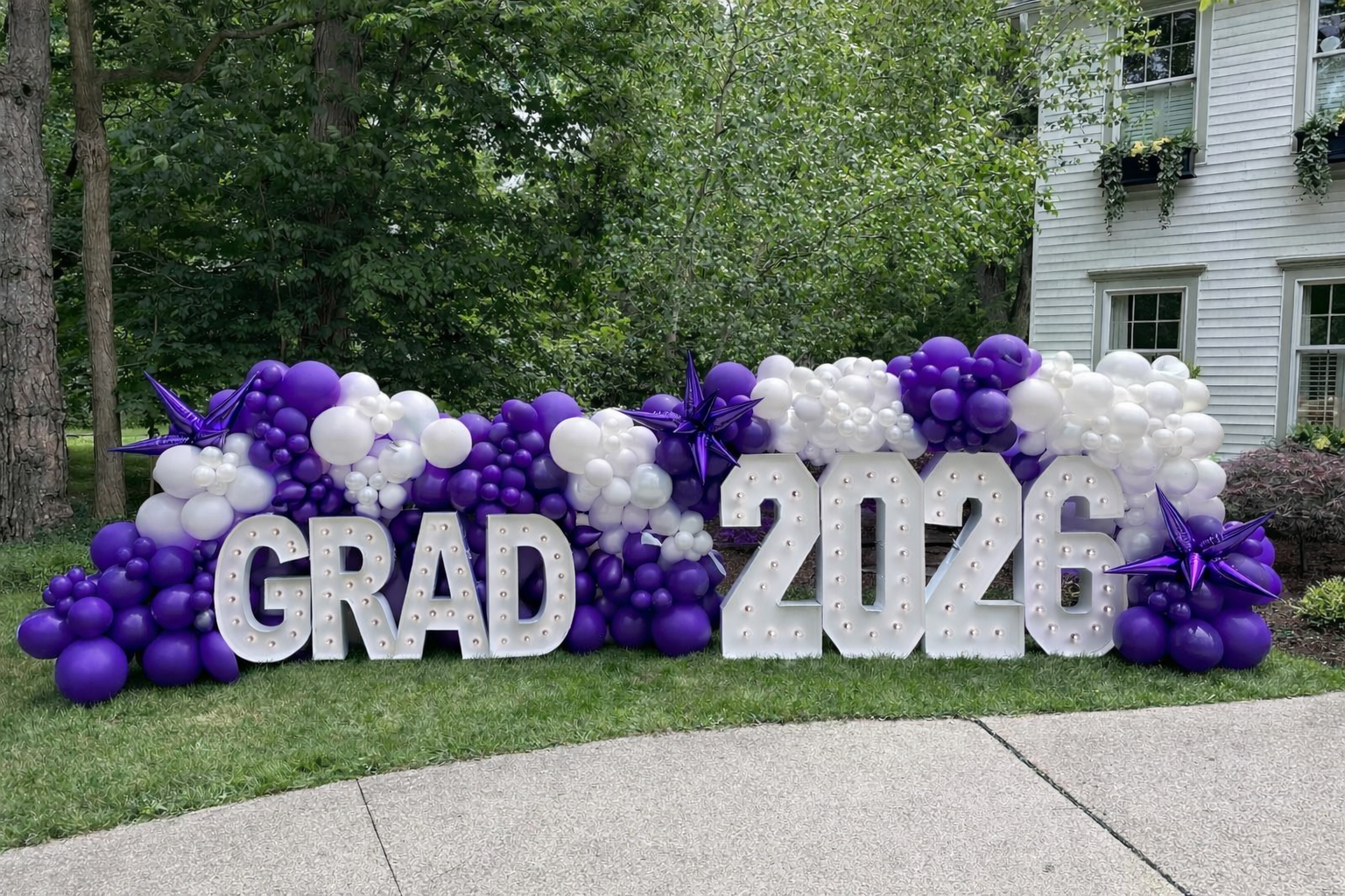 Purple and white 2026 graduation balloon marquee yard display featuring large illuminated 'GRAD 2026' marquee letters, organic purple white balloon garland, chrome purple starburst accents, perfect for high school senior graduation lawn sign, Class of 2026 outdoor photo backdrop, or college grad celebration decor