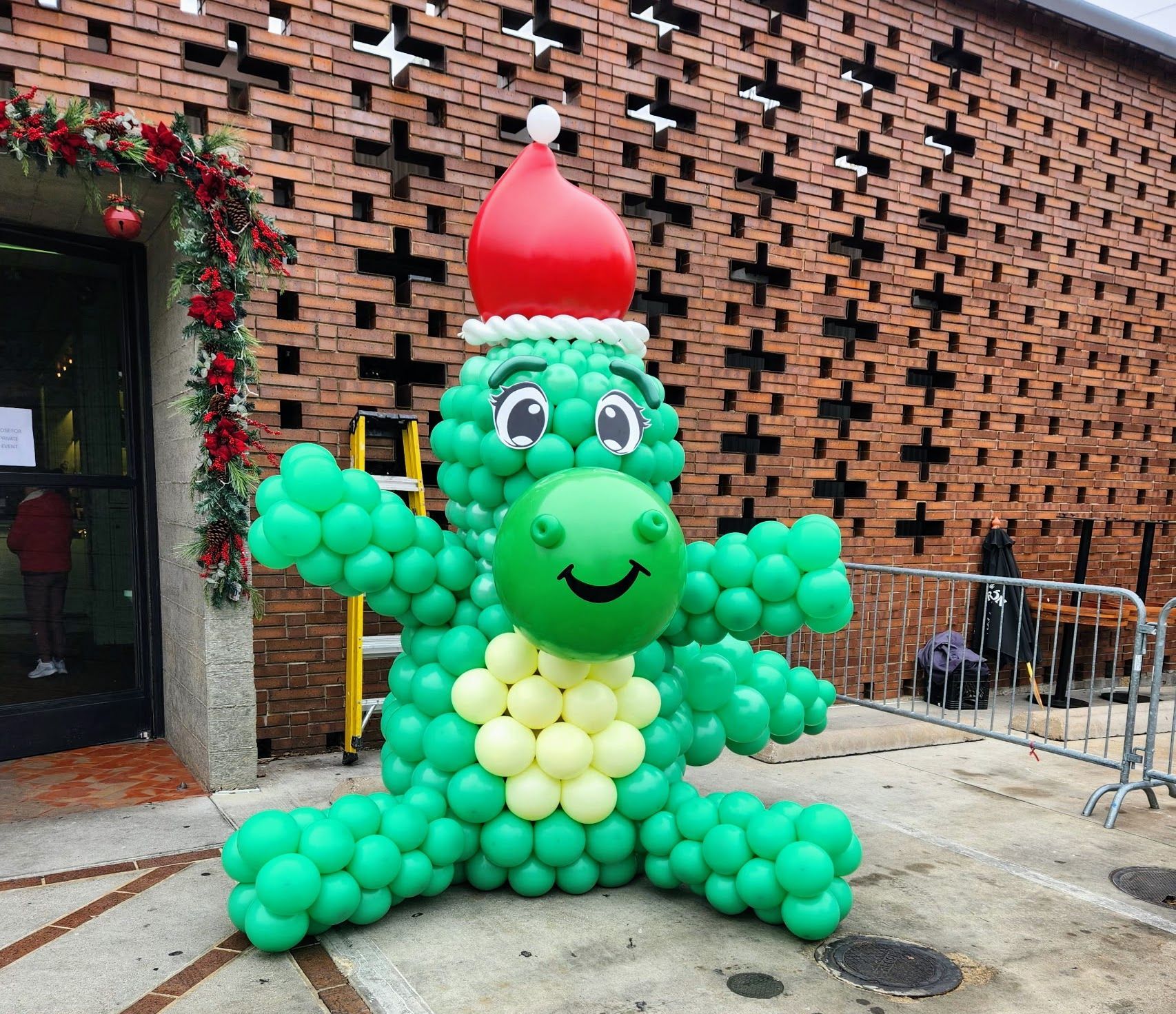 Large balloon sculpture of a happy brontosaurus dinosaur wearing a Santa hat sitting in a parking lot with arms wide open