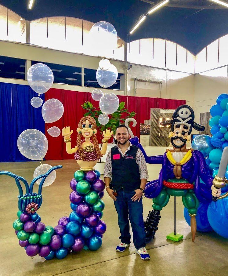 A man is standing in front of a balloon sculpture of a mermaid and a pirate.