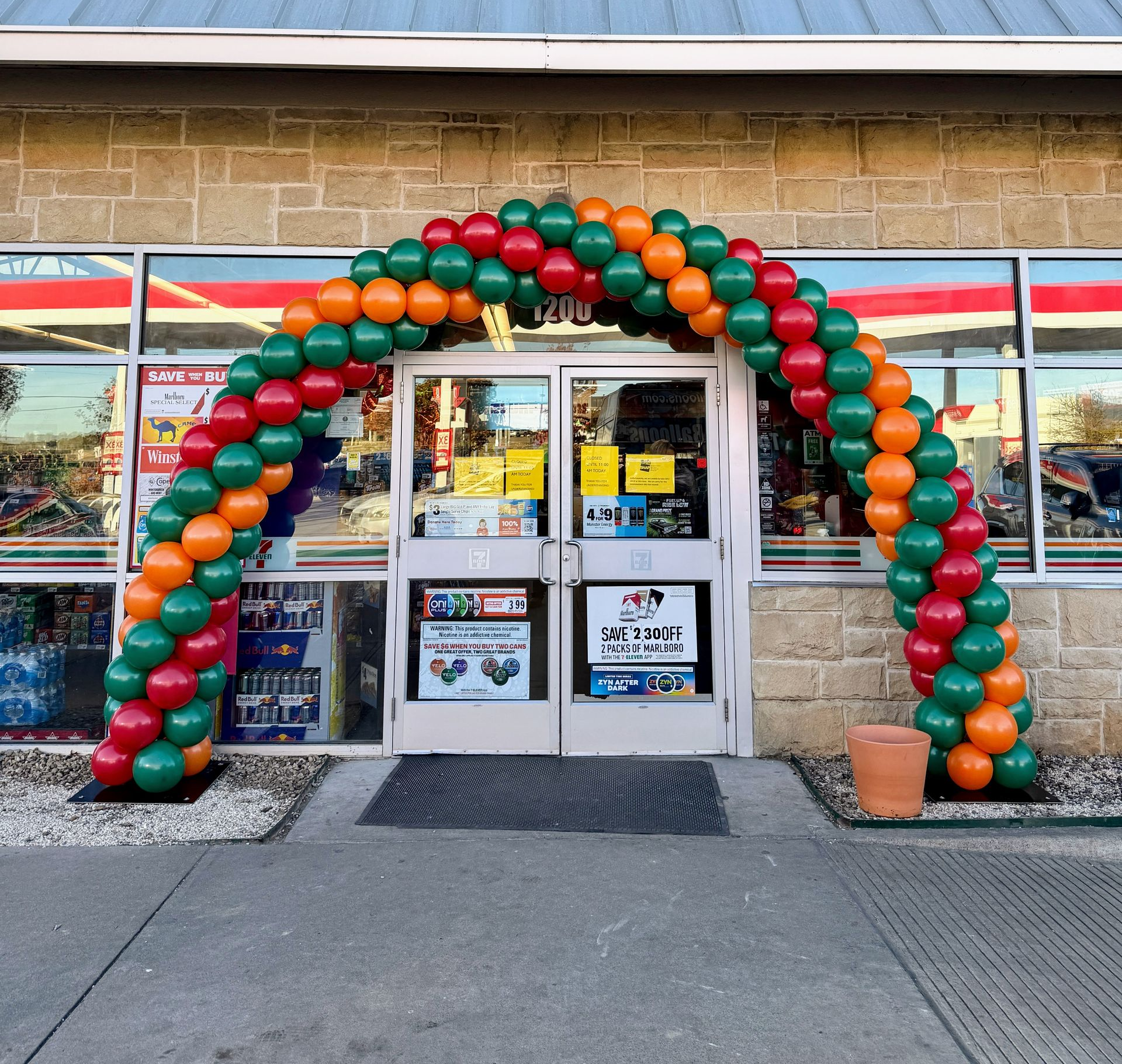 A festive balloon arch in green, orange, and yellow colors adorns the entrance of a 7-Eleven store. This eye-catching decoration enhances the store's facade, making it ideal for grand openings, promotions, or special events. Perfect for retail display ideas, balloon decorations, and attracting customer attention.