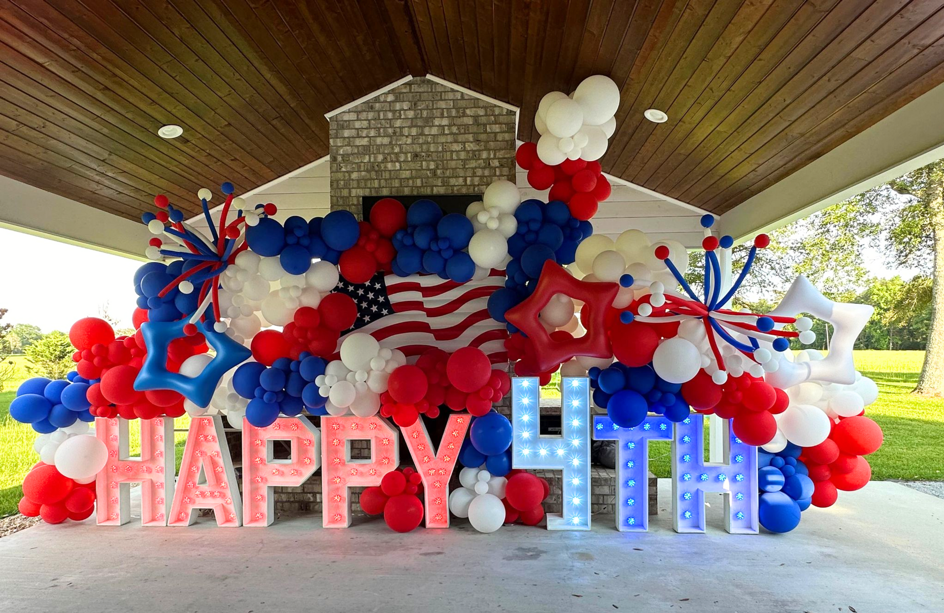Massive patriotic 4th of July balloon installation with red white blue organic garland,  'HAPPY 4TH' marquee letters, American flag backdrop, exploding starburst accents, fireworks clusters, and festive balloon clusters under outdoor pavilion, perfect for Independence Day party, backyard BBQ, or July 4th celebration decor