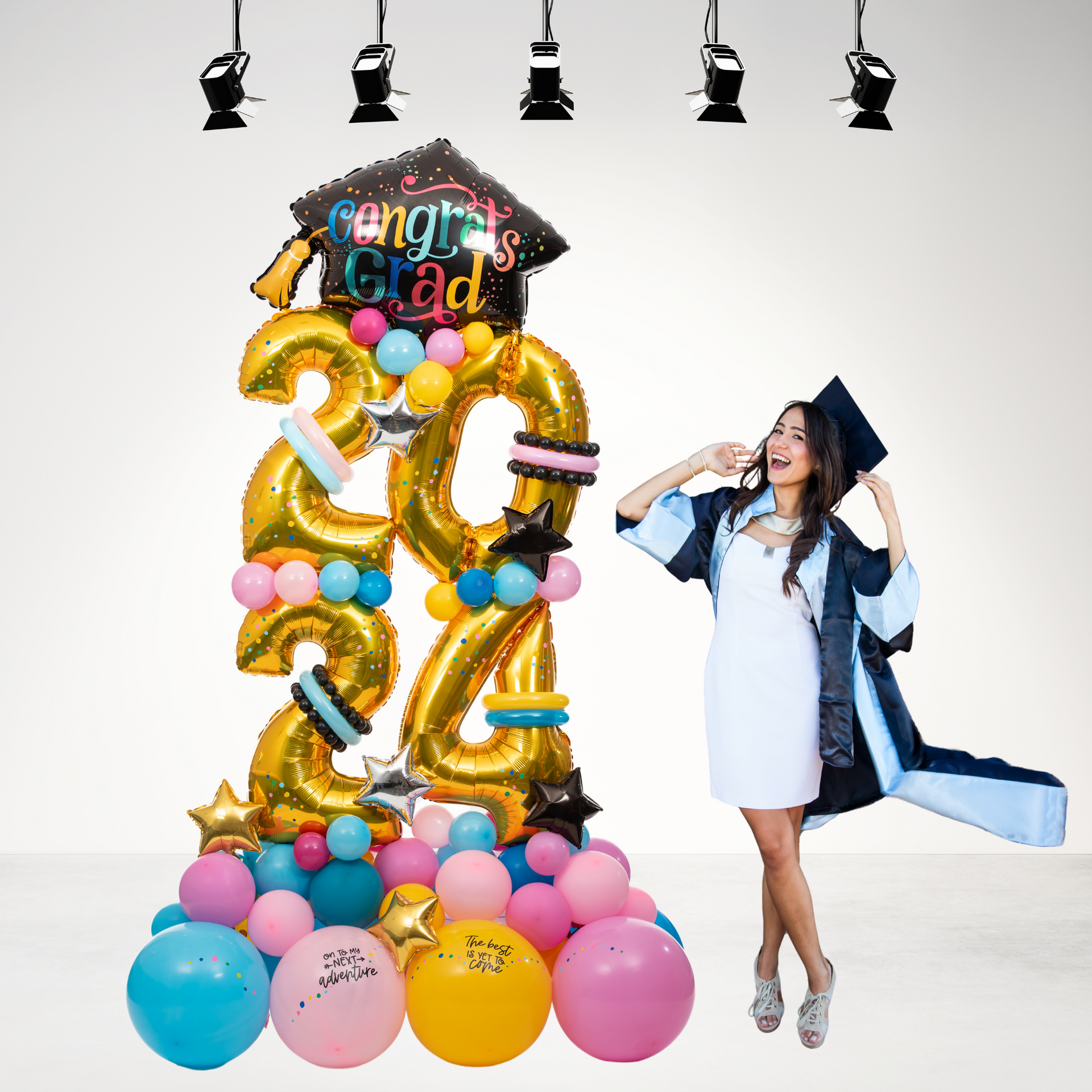 A woman in a graduation cap and gown is standing in front of a congratulations grad balloon