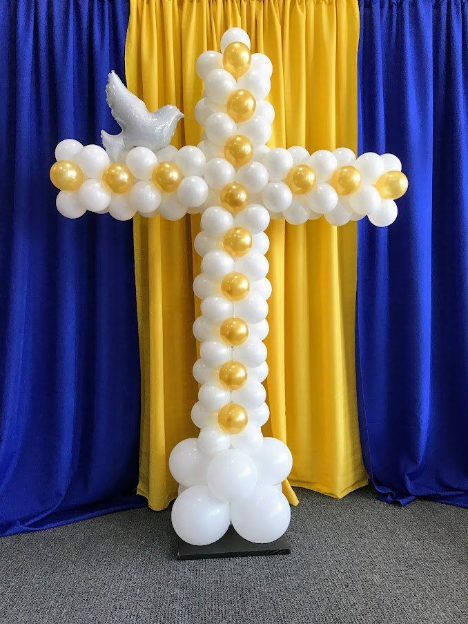 A cross made of balloons with a dove on top is sitting in front of a blue and yellow curtain.