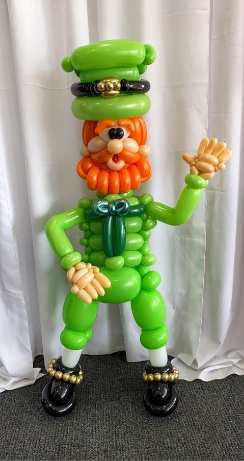A leprechaun made out of balloons is standing in front of a white curtain.