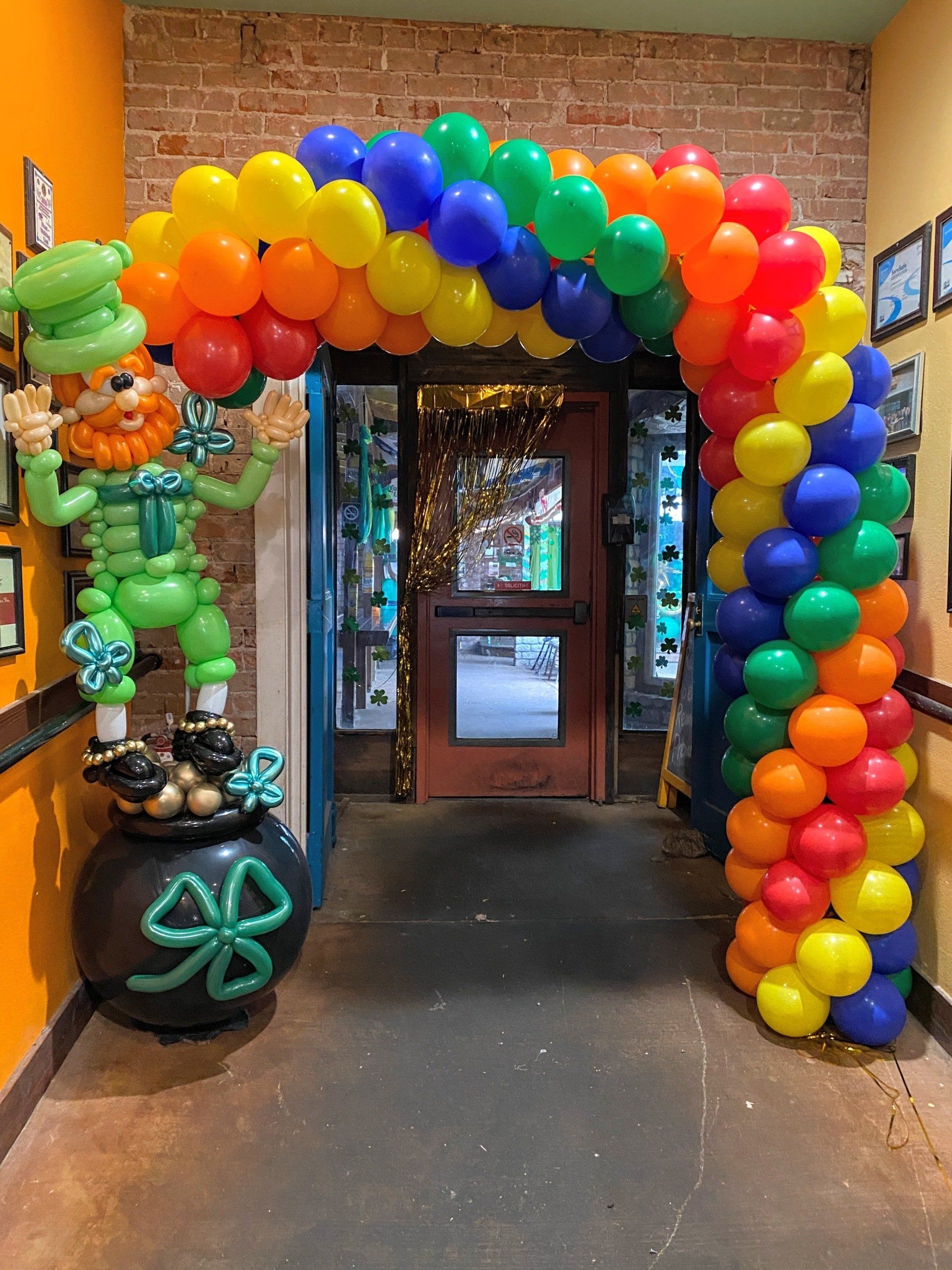 A rainbow colored balloon arch with a leprechaun and a pot of gold.