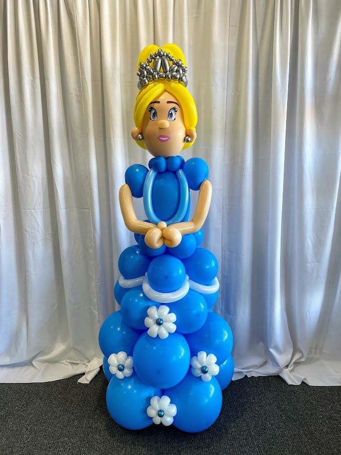 A princess made out of blue balloons is standing in front of a white curtain.