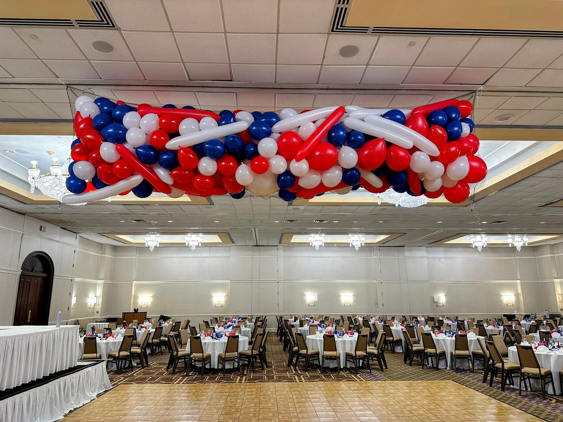 A red white and blue balloon drop suspended from ballroom ceiling, featuring dramatic organic garland with red blue white latex balloons and twisted accents, perfect for grand opening, graduation ceremony, product launch, corporate event or patriotic celebration balloon drop
