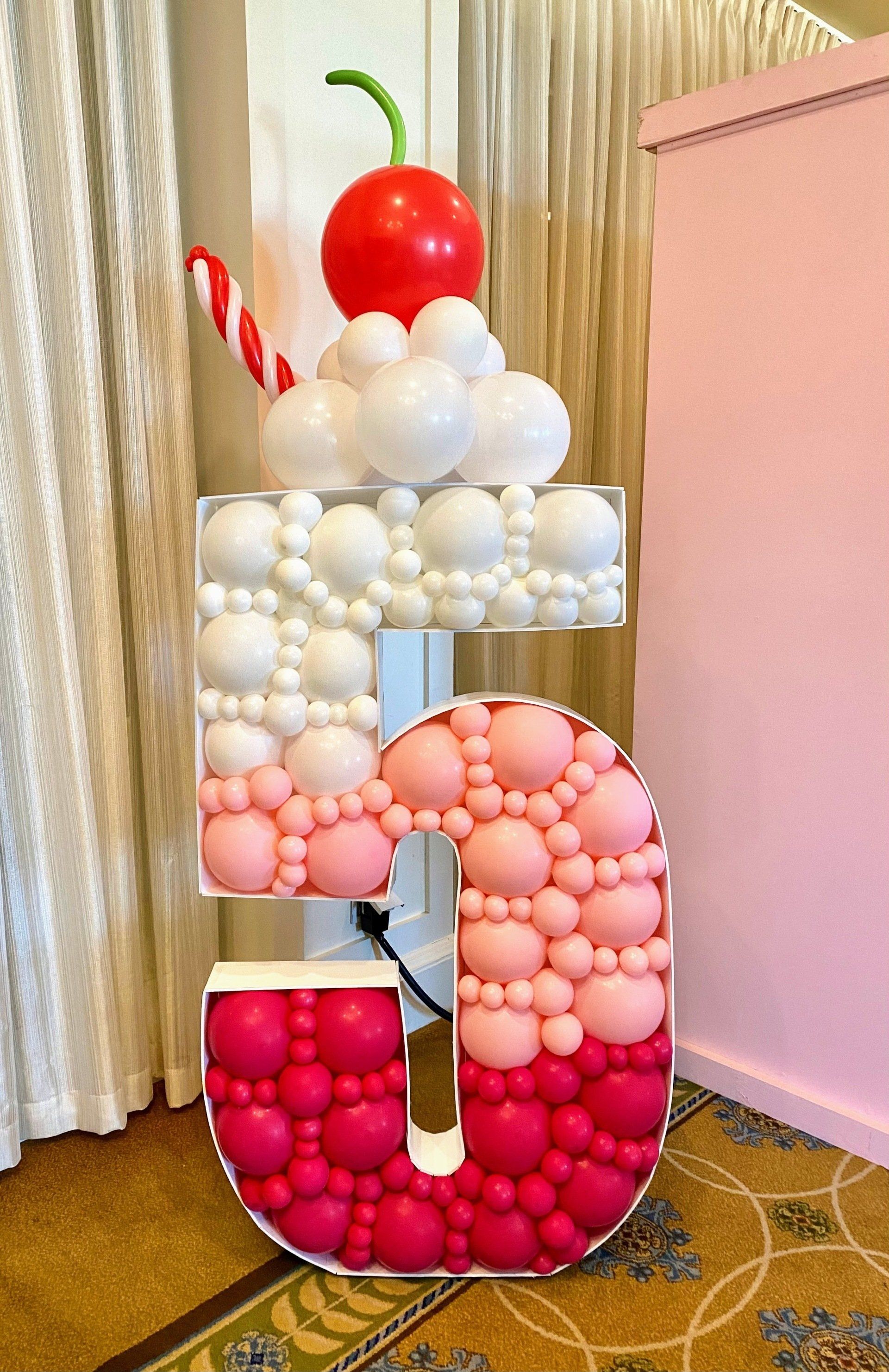 A large number 5 made of pink and white balloons with a cherry on top.