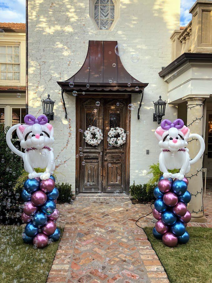 Two balloon cats are sitting on top of balloon columns in front of a house.