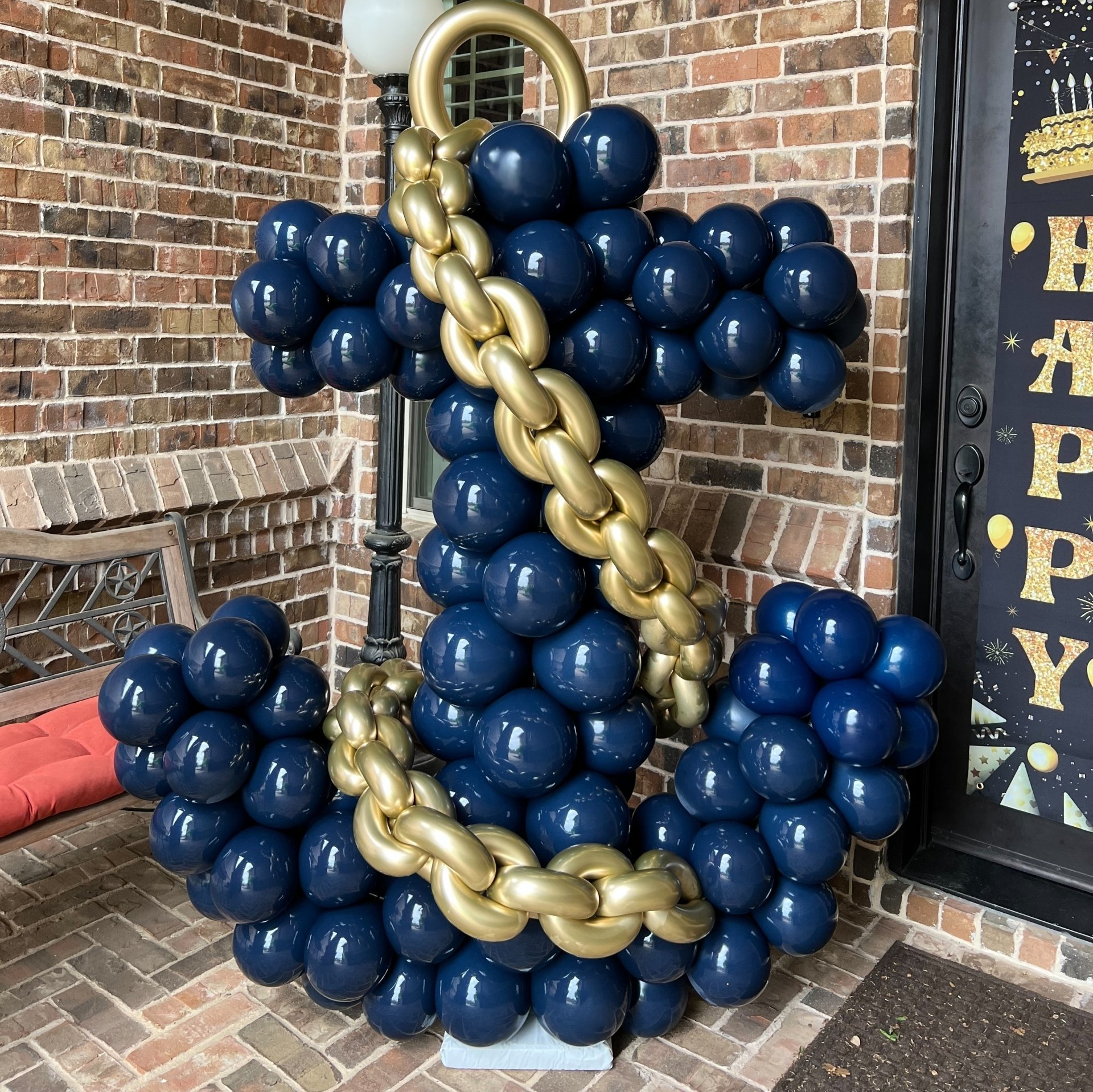 A large anchor made of blue and gold balloons is sitting on a brick porch.