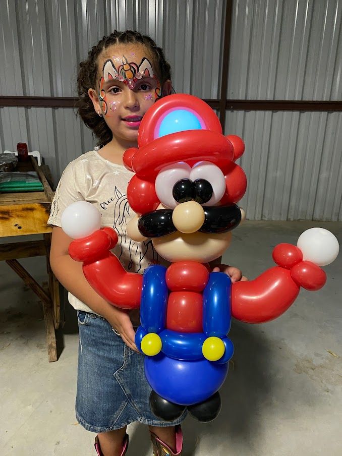 A little girl with face paint is holding a mario balloon.