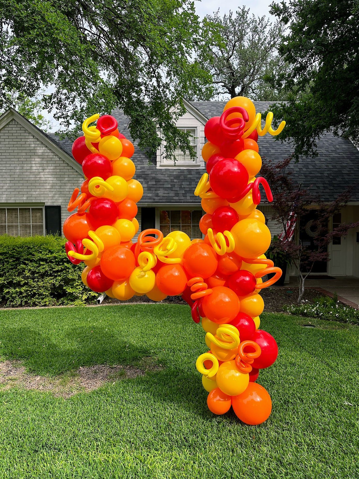 A letter u made of orange and yellow balloons is sitting in the grass in front of a house.