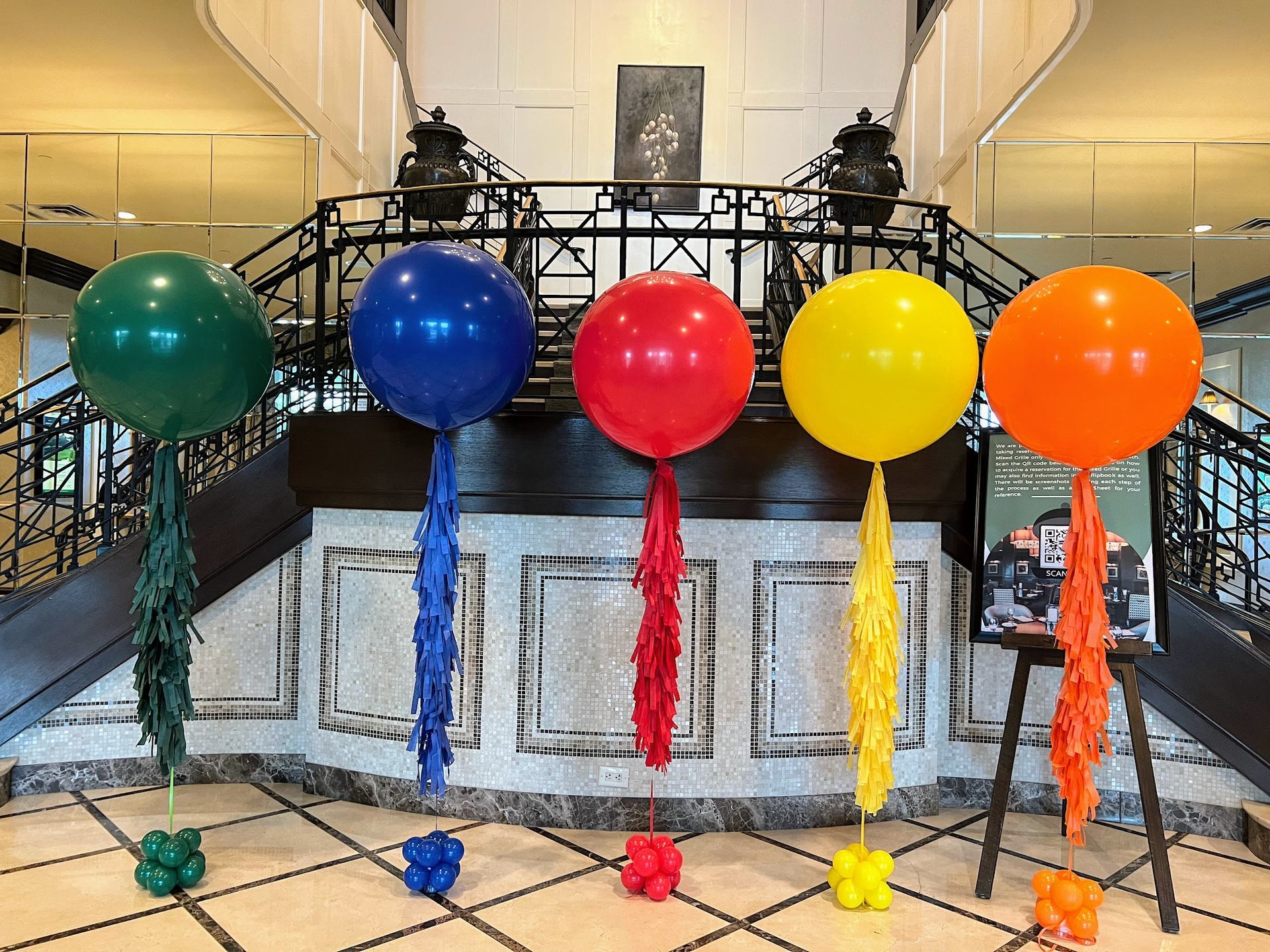 Grand hotel lobby staircase decorated with jumbo colorful helium balloons in green, blue, red, yellow, and orange, each topped with matching long fringe tassels, perfect for event entrance, corporate celebration, or festive party decor