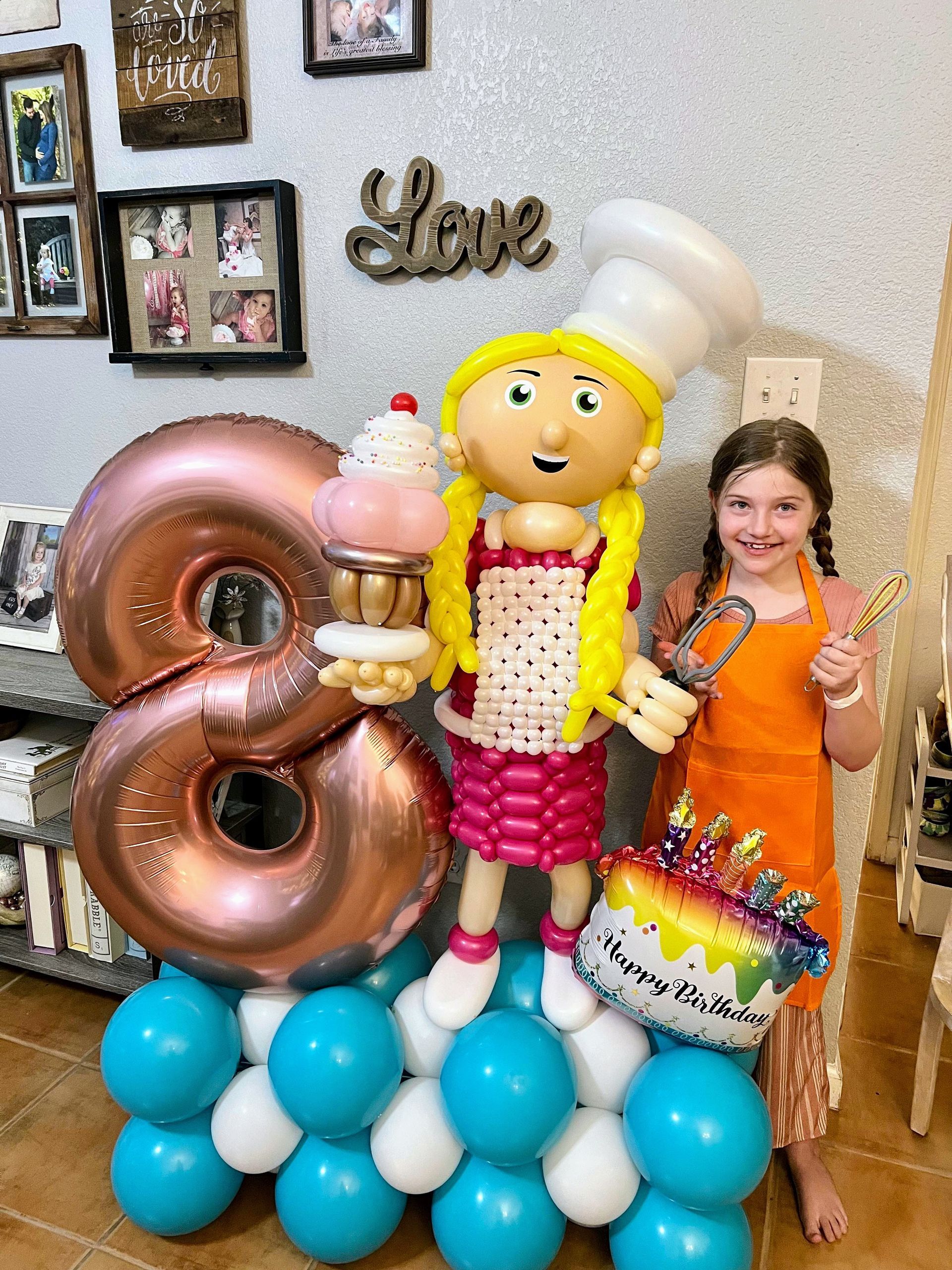 A little girl is standing next to a balloon figure of a girl holding a cupcake.