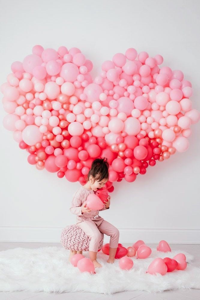 A little girl is sitting in front of a heart made of pink balloons.