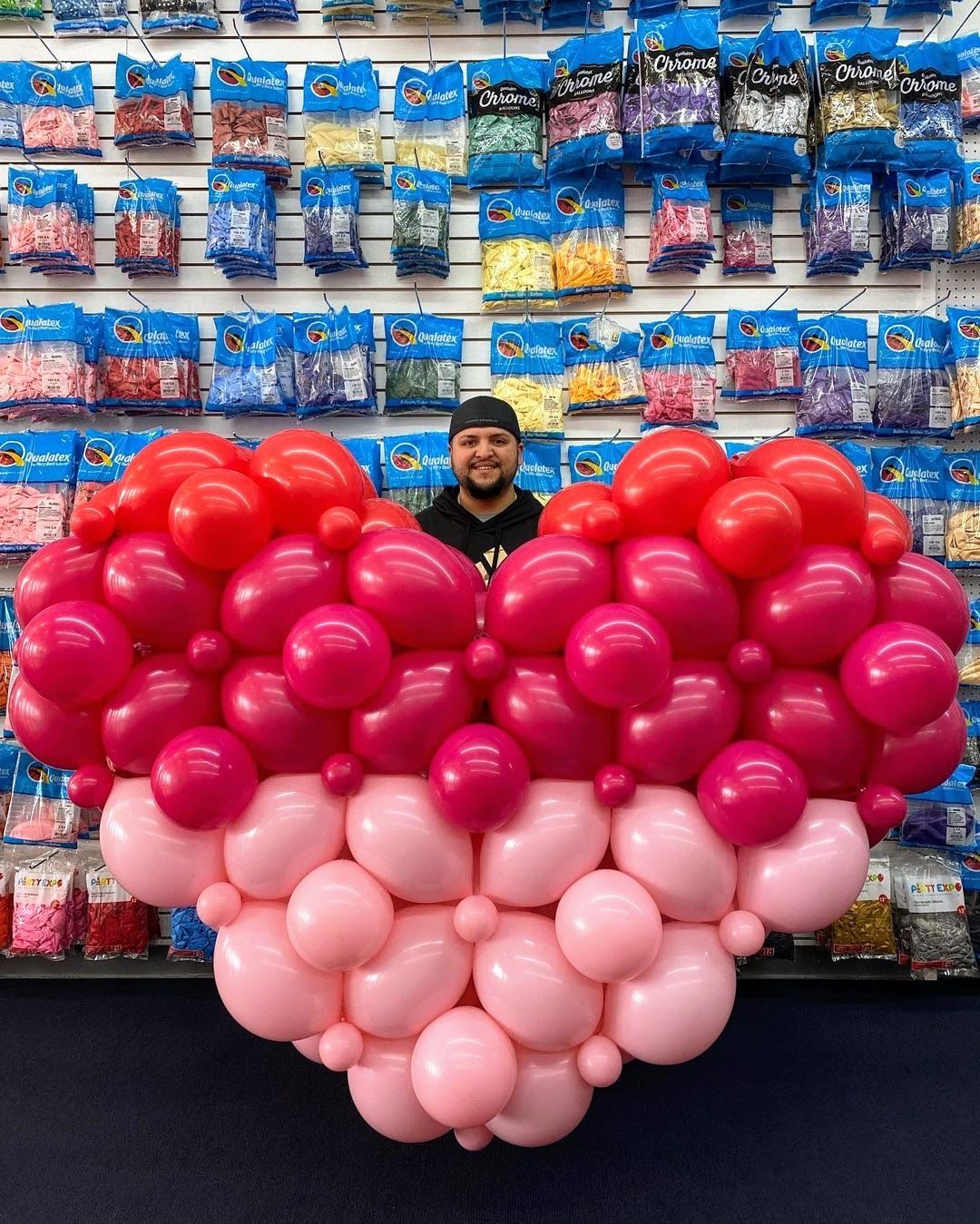 A man is standing in front of a large heart made of pink and red balloons.
