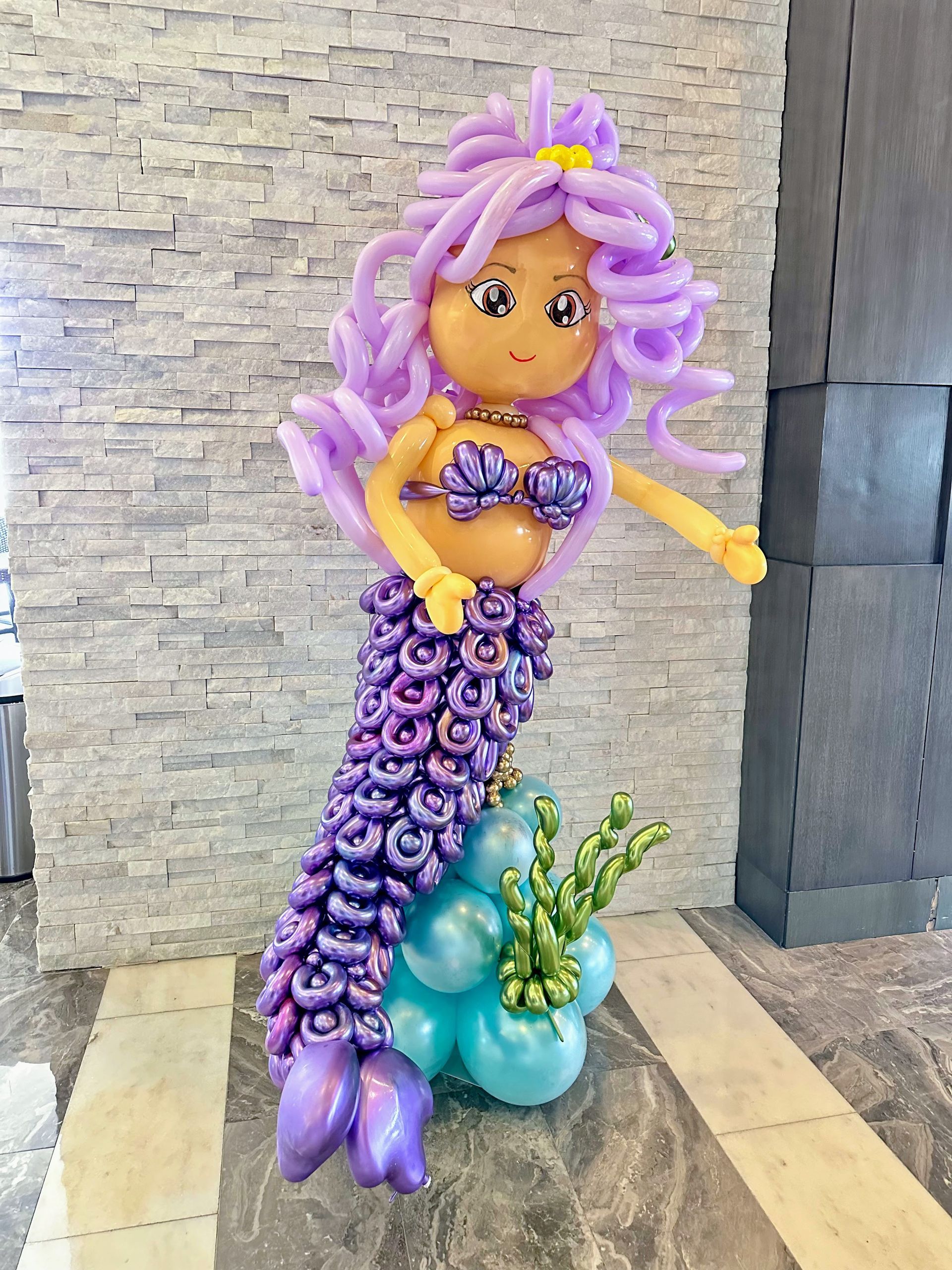 A mermaid made out of balloons is standing in front of a brick wall.