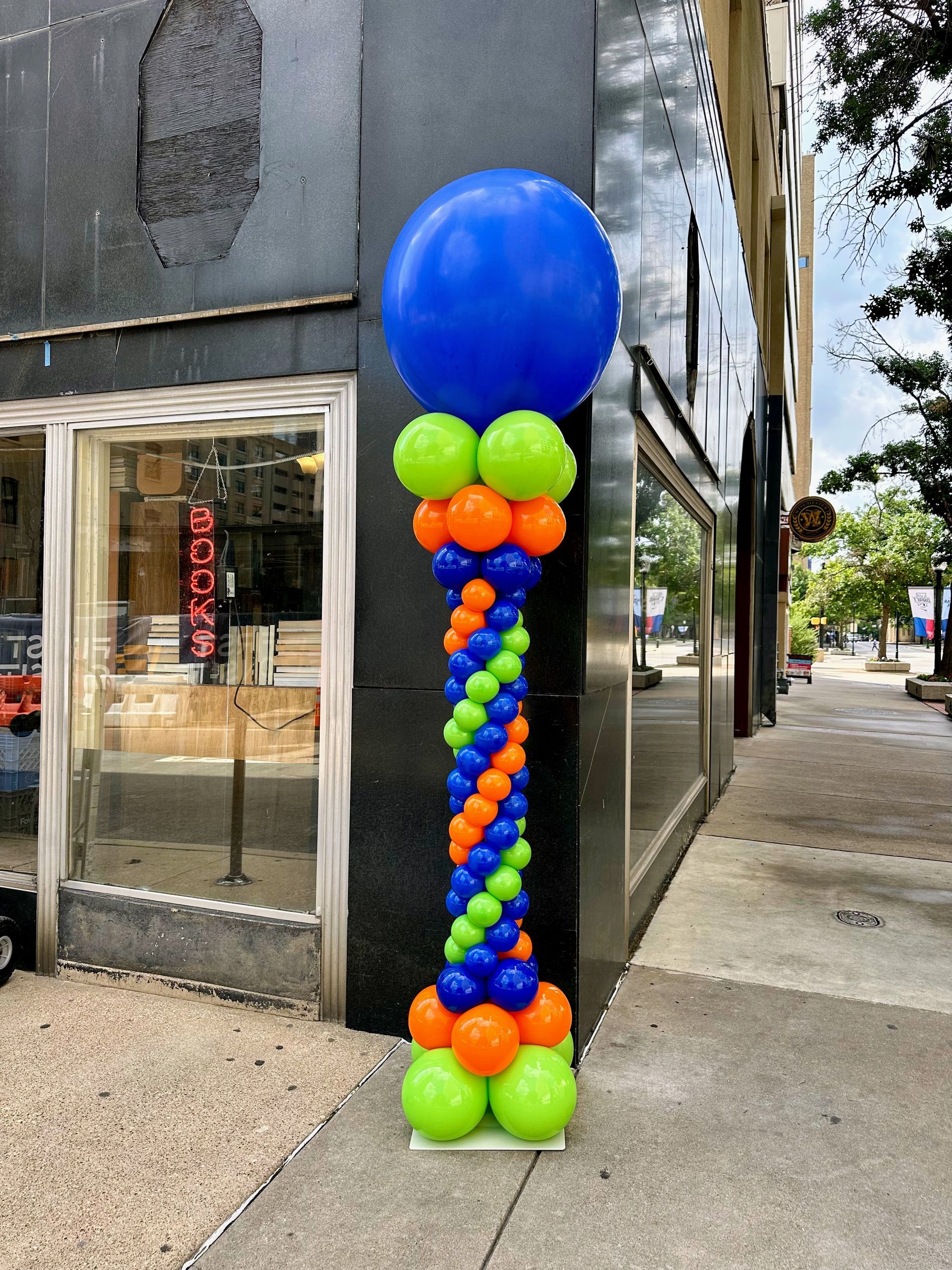 A colorful balloon column featuring a large blue balloon at the top, surrounded by vibrant green, orange, and blue balloons arranged in a spiral pattern. This eye-catching display enhances the storefront, making it perfect for grand openings, events, or promotions. Ideal for retail decorations, balloon artistry, and attracting customer attention.