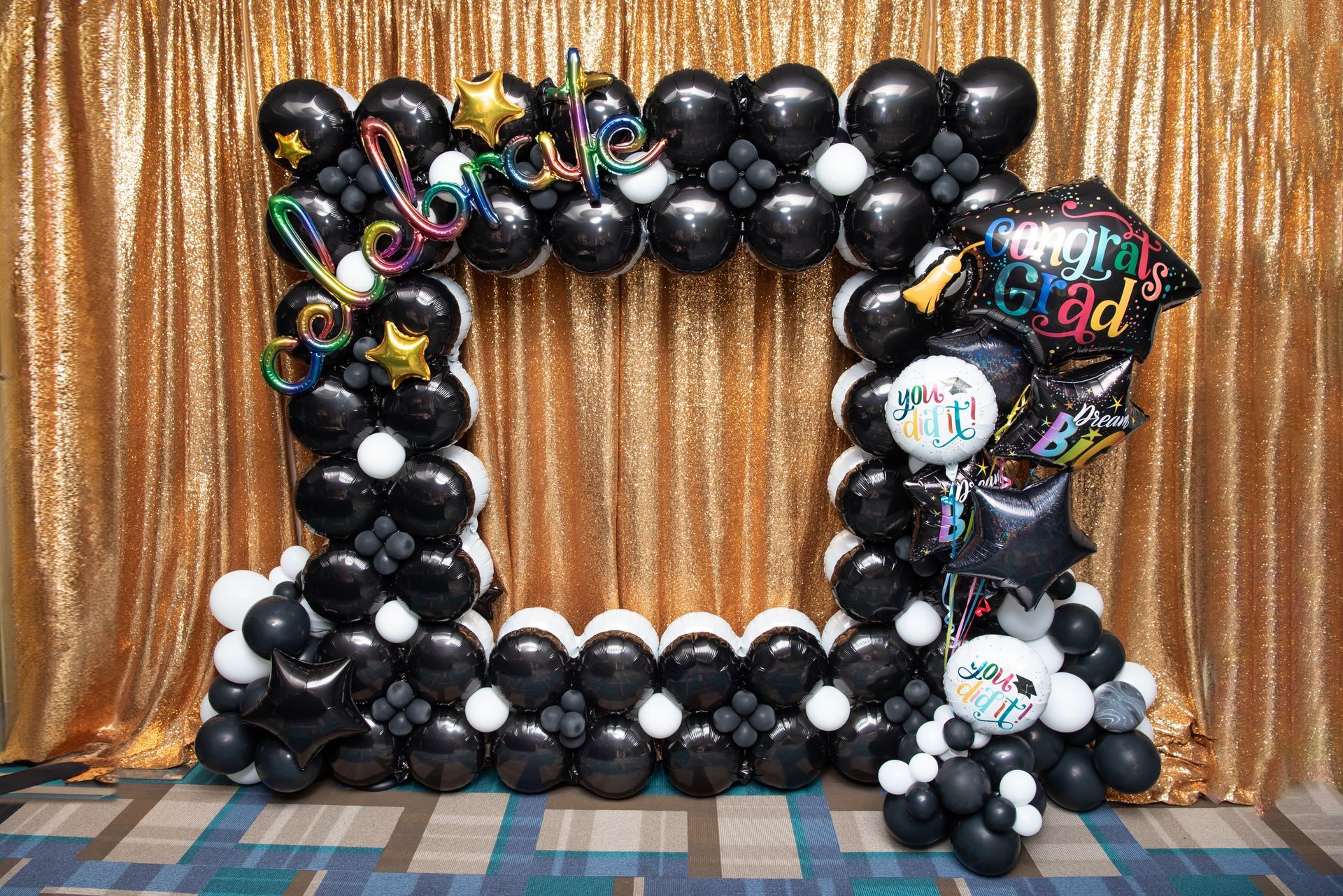 Stunning black foil graduation photo booth frame with iridescent rainbow chrome 'Congrats Grad' foil letters, metallic gold stars, 'You Did It' and grad cap foil balloons, black chrome latex base on shimmering gold curtain backdrop, perfect for high school or college graduation pictures and celebrations