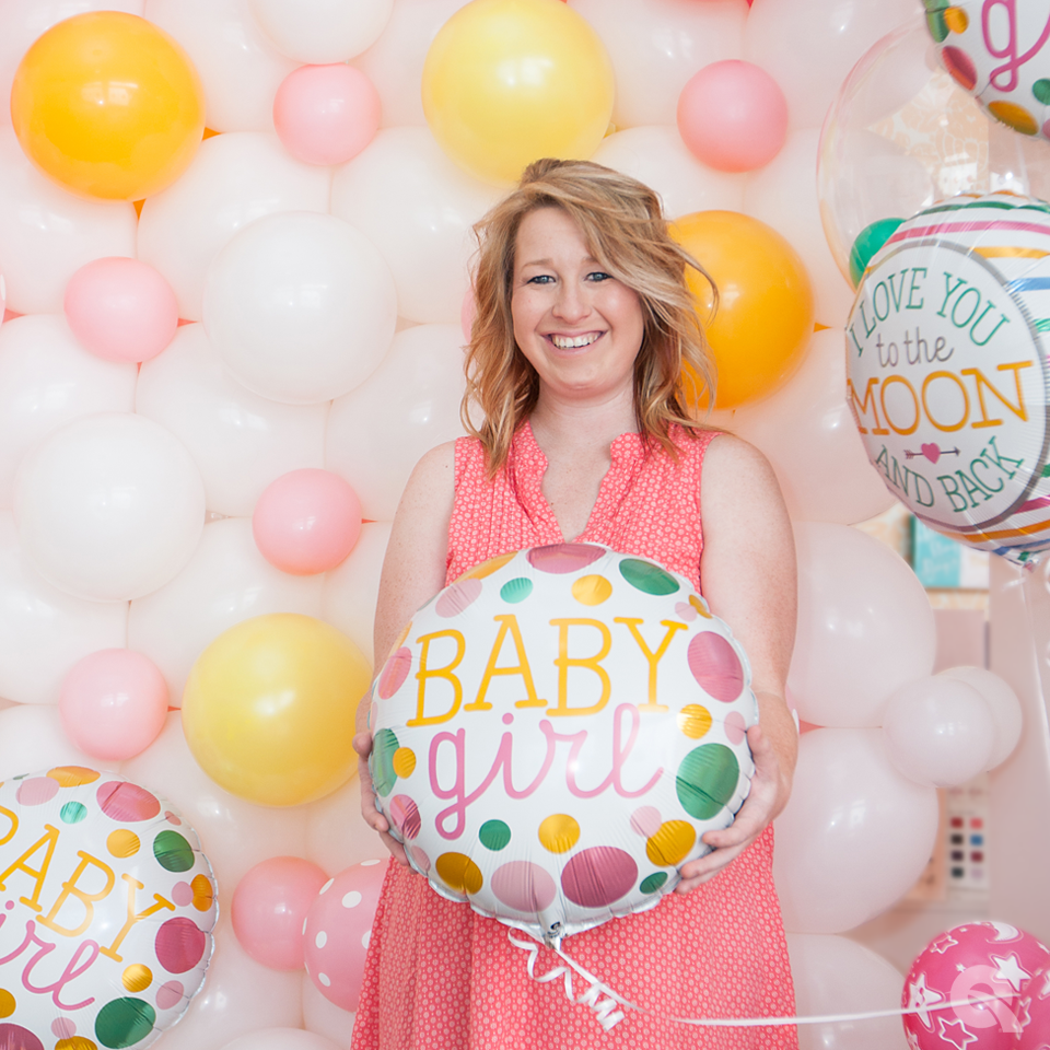 a woman holding a baby girl balloon in front of a wall of balloons