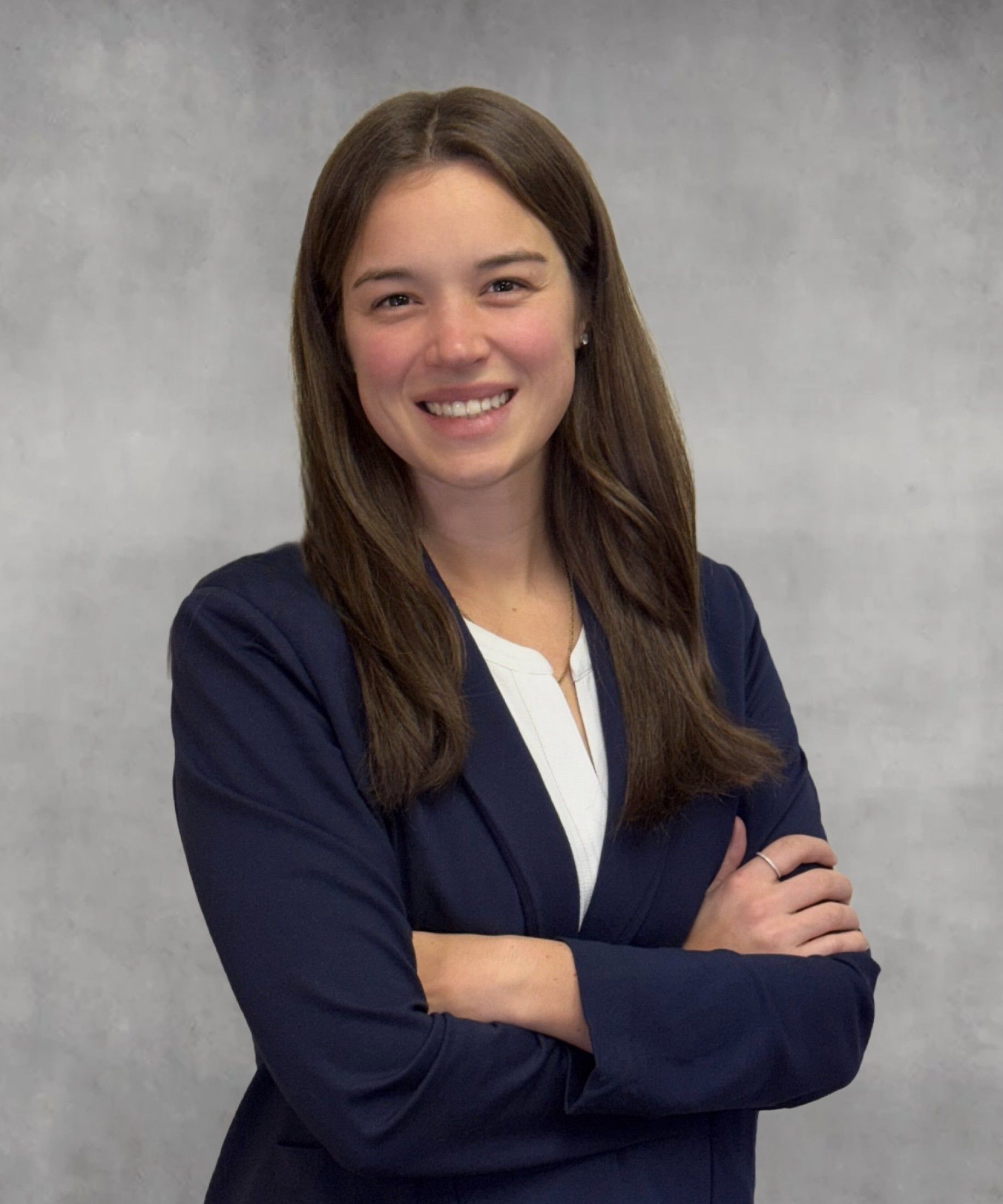 Woman in navy blazer smiles, arms crossed, white shirt, against a gray wall.