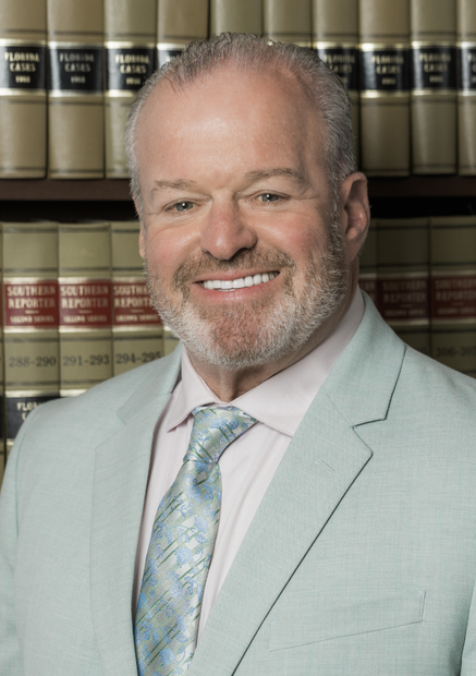 Smiling man in a light gray suit and patterned tie standing before a bookshelf of law books