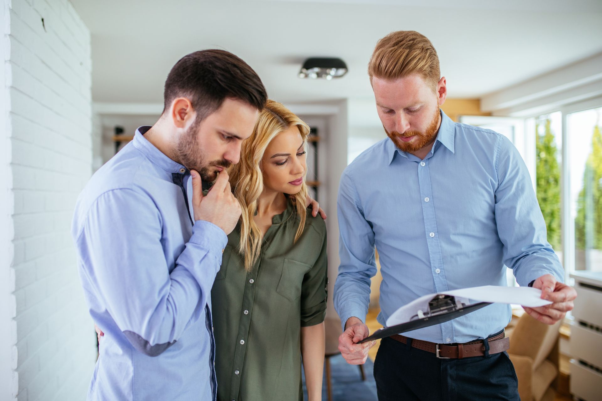A man and a woman look thoughtfully at a document held by a professional in a light-filled office space.
