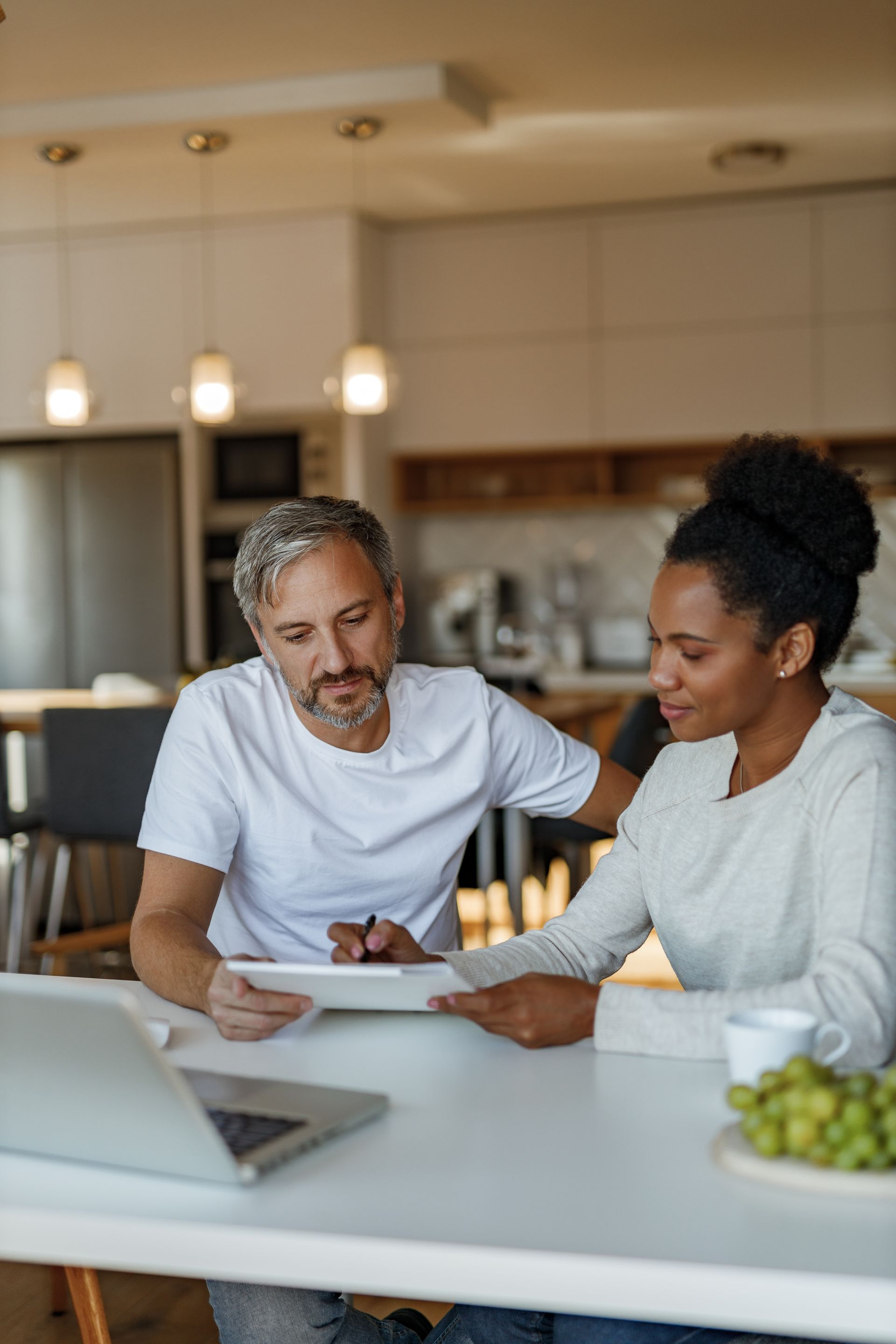 Two people collaborate on documents at a kitchen table with a laptop and a bowl of grapes nearby.