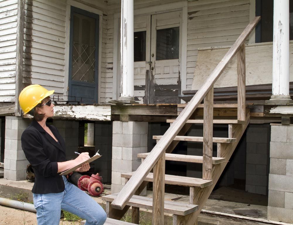 A Woman Wearing A Hard Hat Is Writing On A Clipboard In Front Of A House — Salt Building & Pest Inspections in Mudgeeraba, QLD