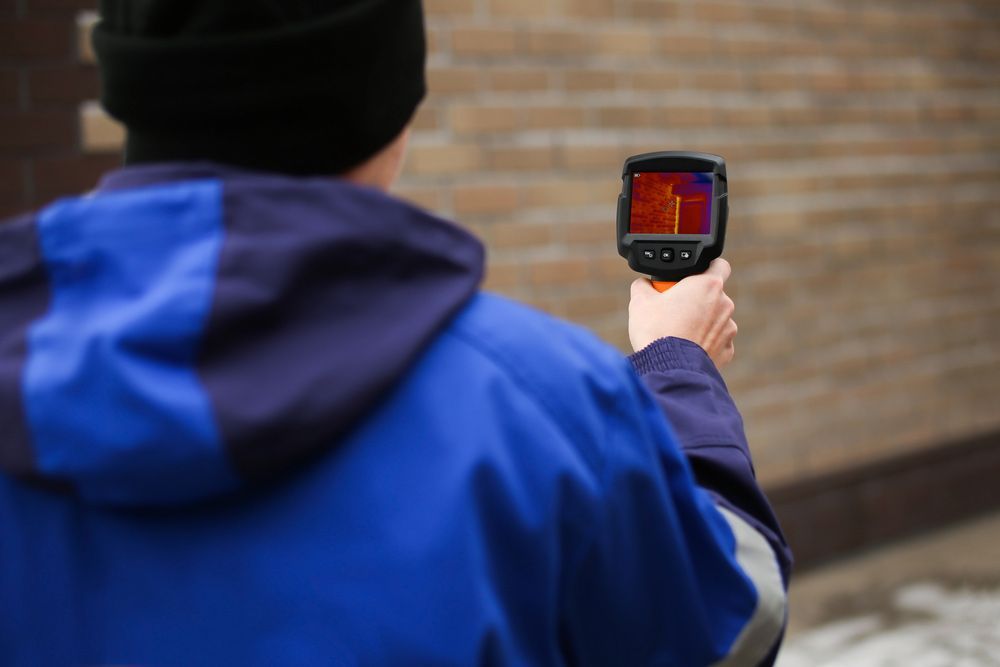 A Man Is Holding A Thermal Camera In Front Of A Brick Wall — Salt Building & Pest Inspections in Varsity Lakes, QLD