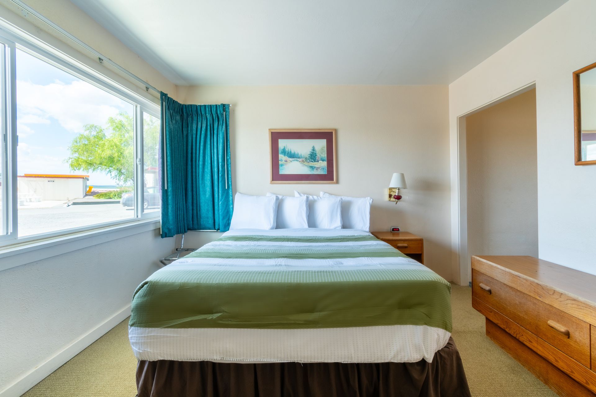 A hotel room with a bed covered in green and white bedding, a large window with teal curtains, and a wooden dresser.