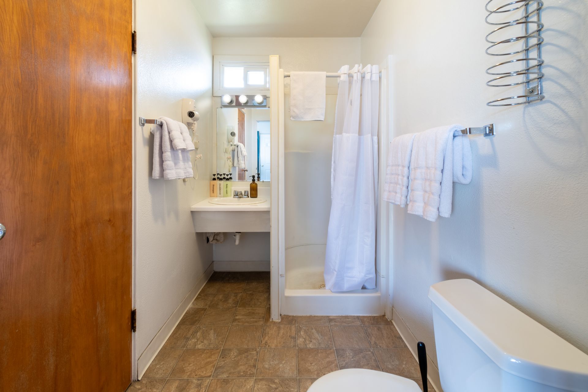 A small bathroom with a white shower, sink, and toilet. A brown door stands on the left, and a towel rack is on the right.