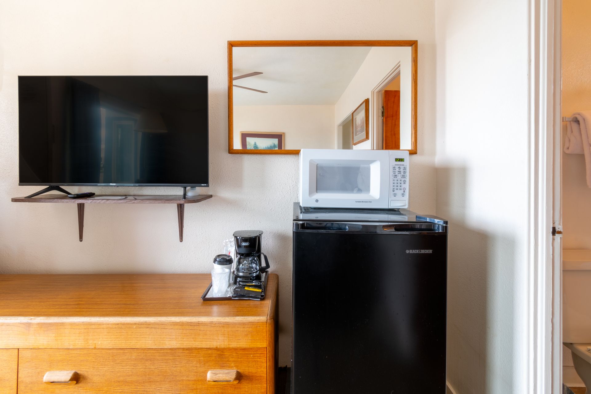 A hotel room setup with a TV, mirror, microwave, mini-fridge, and coffee maker. The door to the bathroom is on the right.