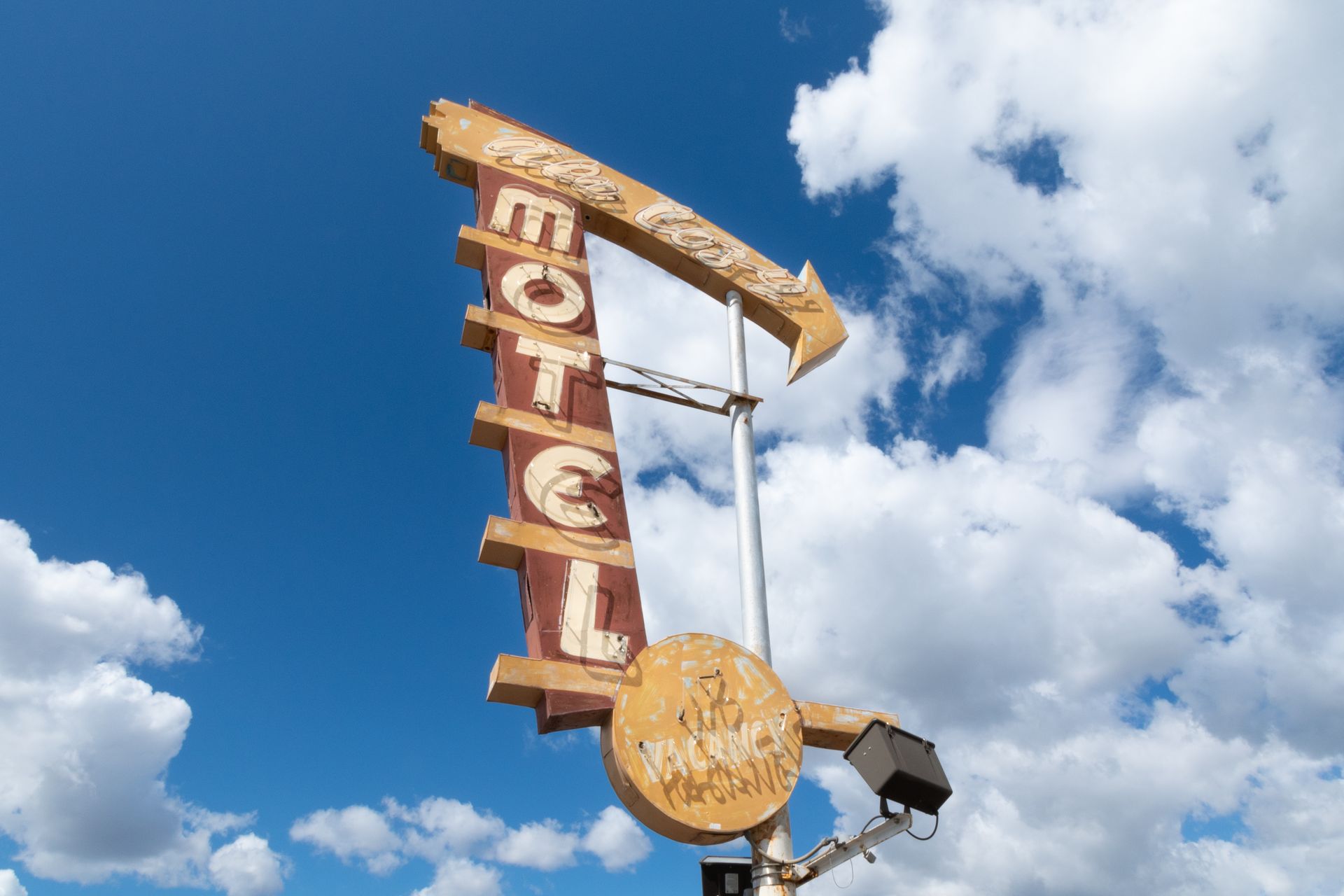 Weathered motel sign with "MOTEL" illuminated, pointing toward the sky; against a bright blue sky with puffy white clouds.