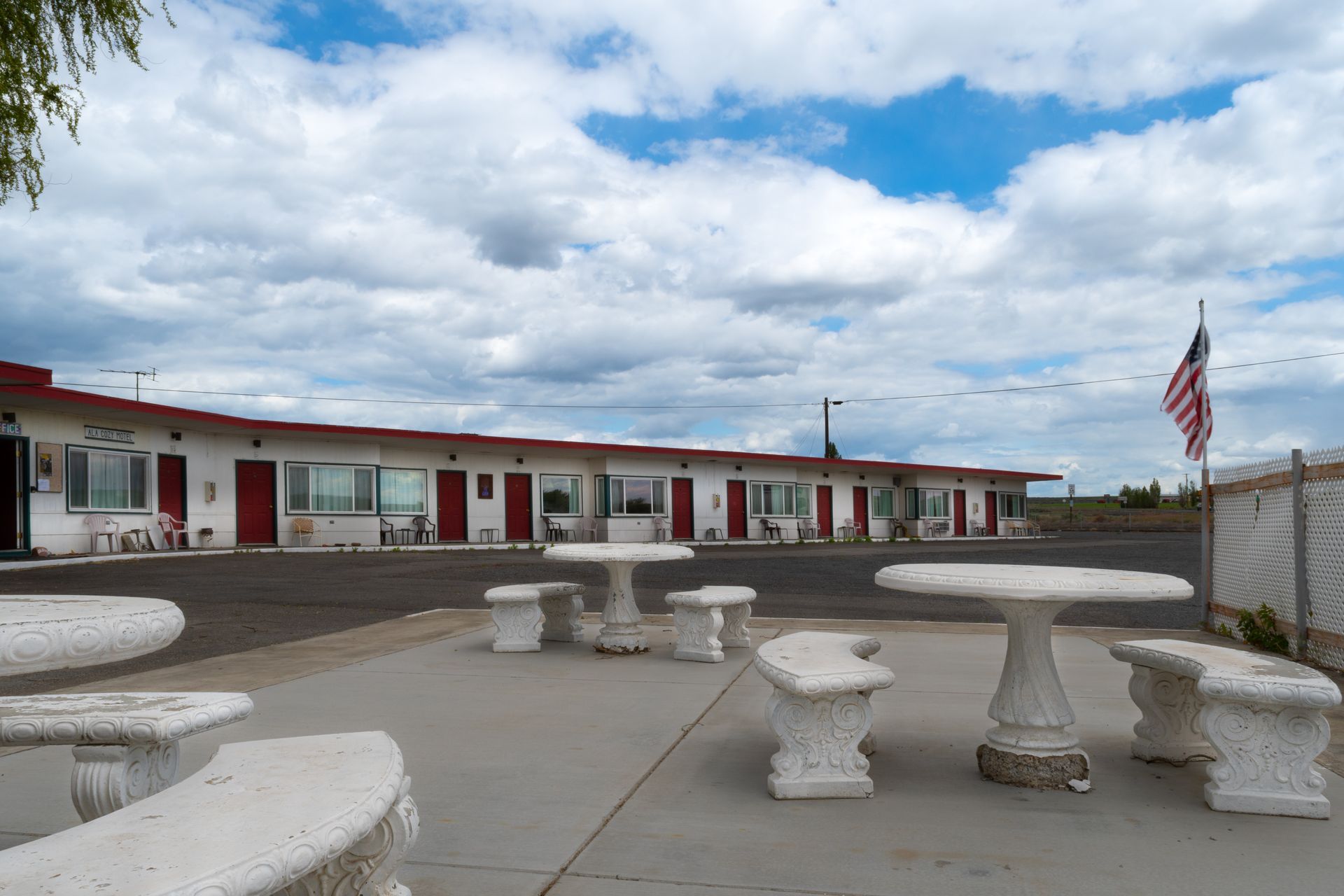 A roadside motel with a row of red doors and white tables. An American flag waves under a cloudy sky.