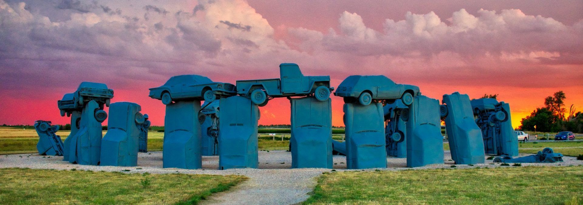 A blue carhenge sculpture against a colorful sunset sky in a field.