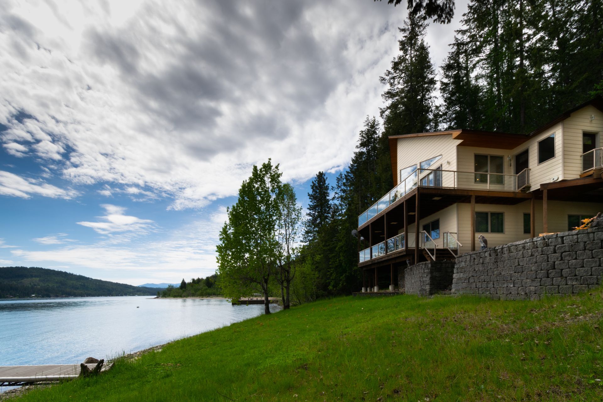 A multi-story house with balconies overlooks a lake. Green grass slopes down to a dock, with a cloudy sky in the background.