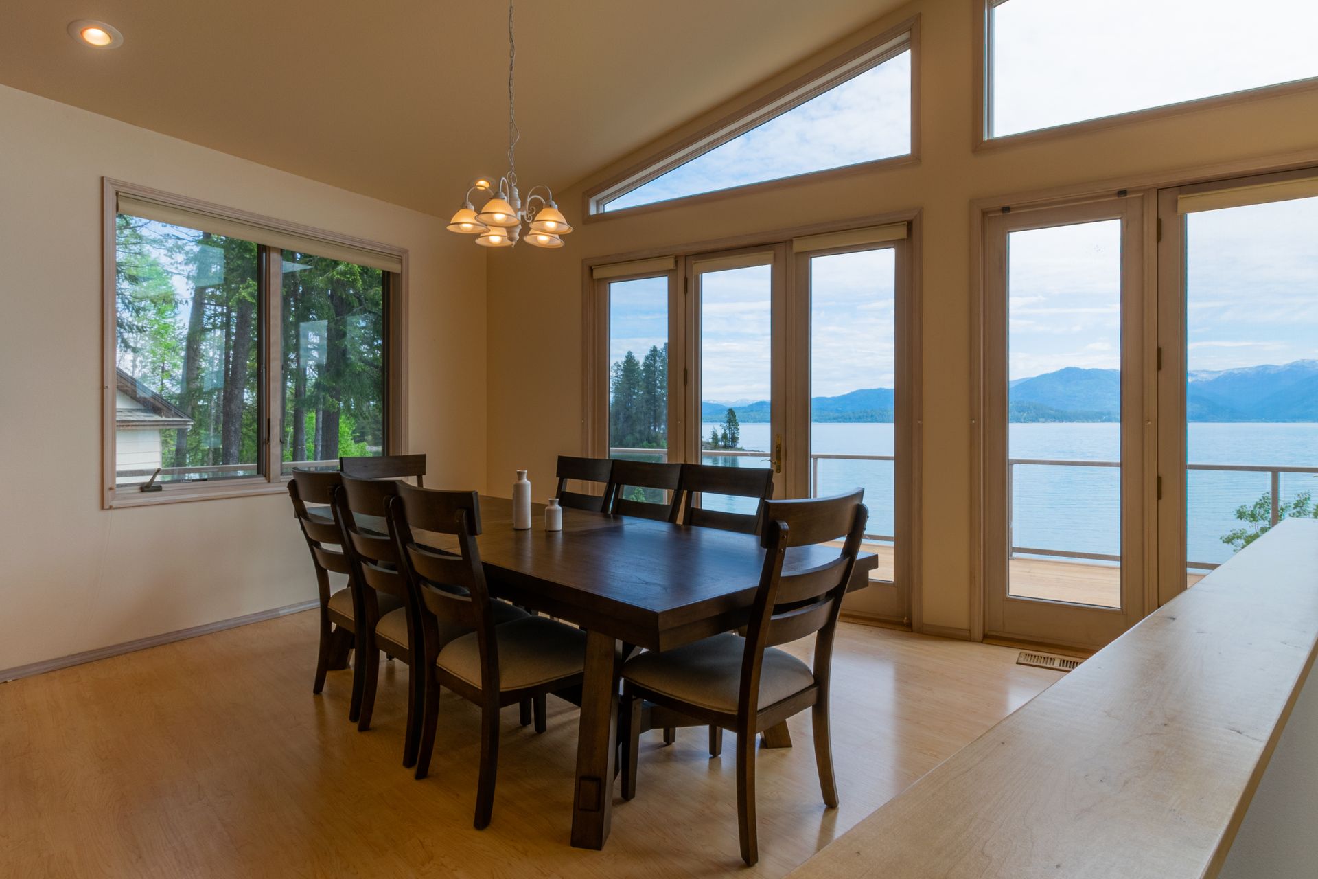Dining room with a large wooden table and chairs, overlooking a lake through large windows.