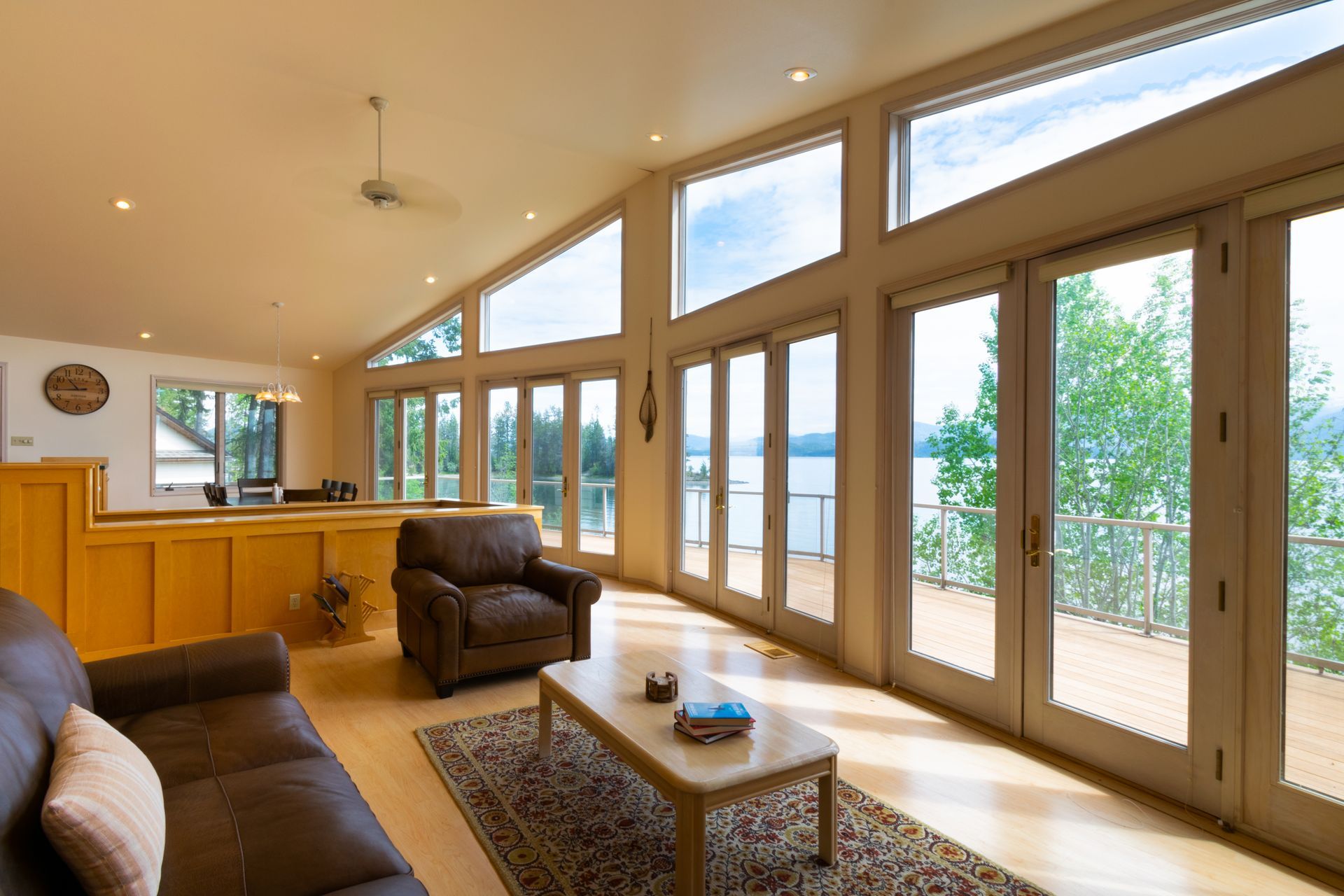 Bright living room with a leather sofa, armchair, and coffee table. Large windows overlook a lake and deck.