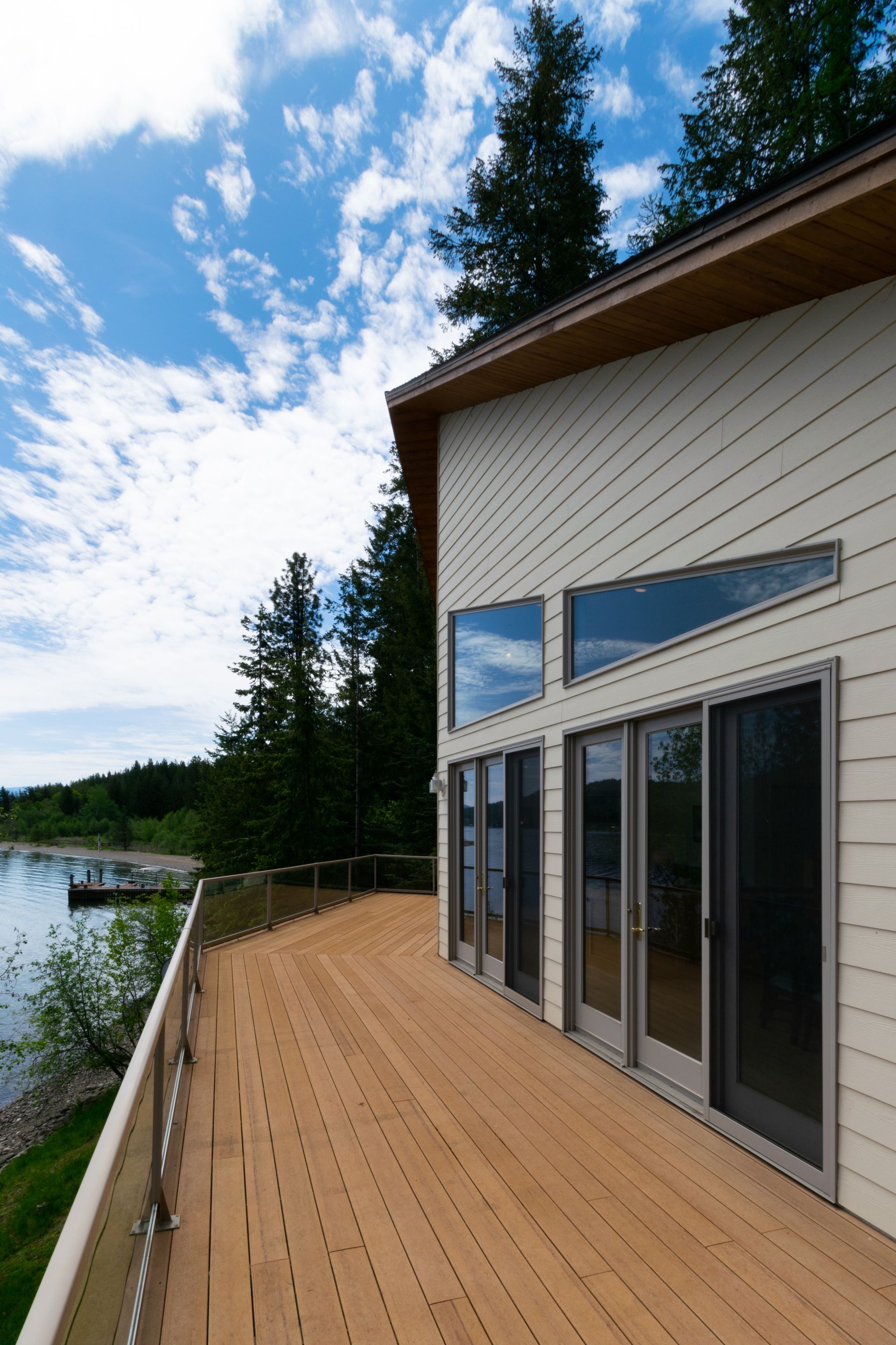 Wooden deck overlooking a lake, beside a modern white house with large windows. Blue sky and trees in the background.