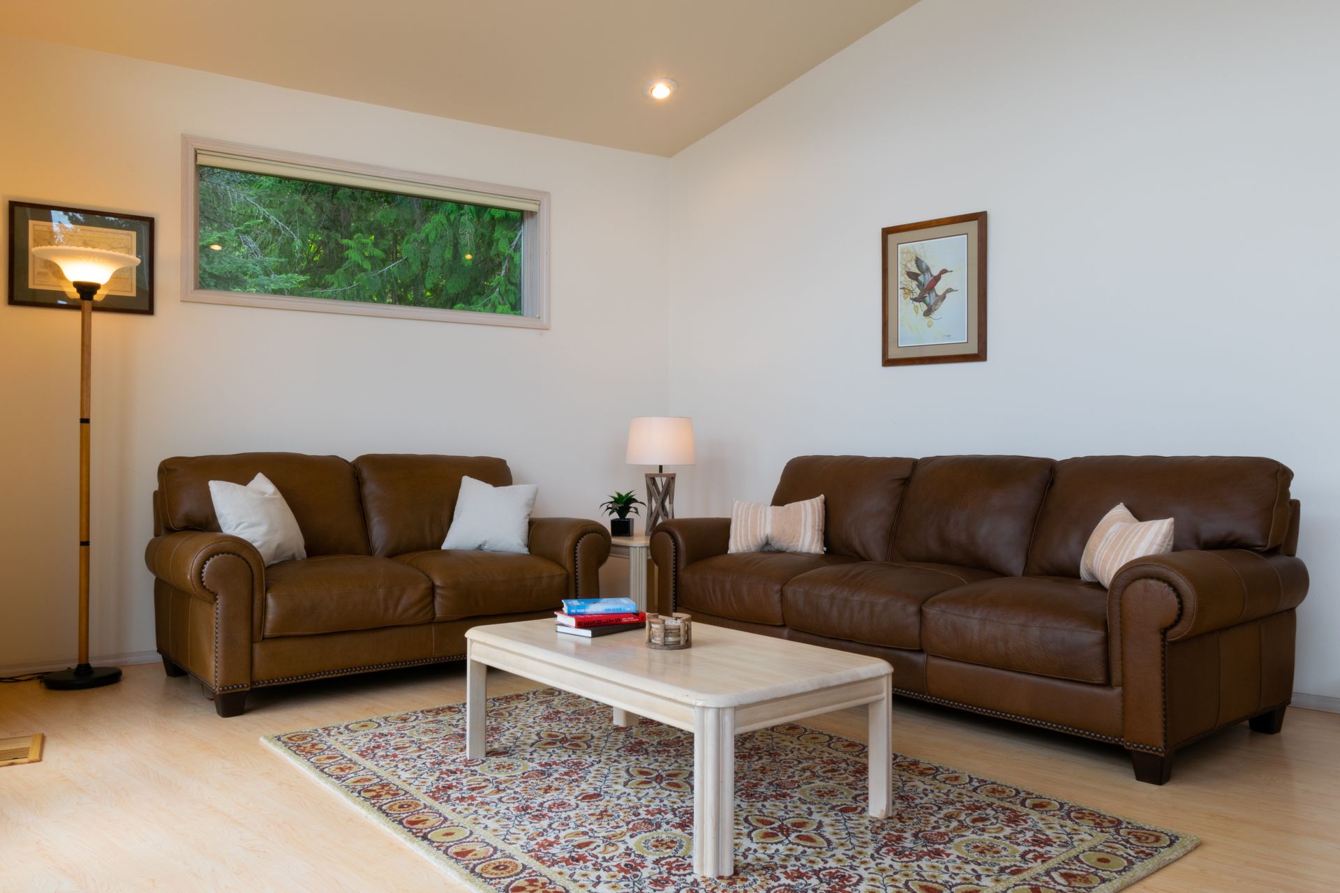 A living room with brown leather sofas, a coffee table, and a rug. A window overlooks greenery.