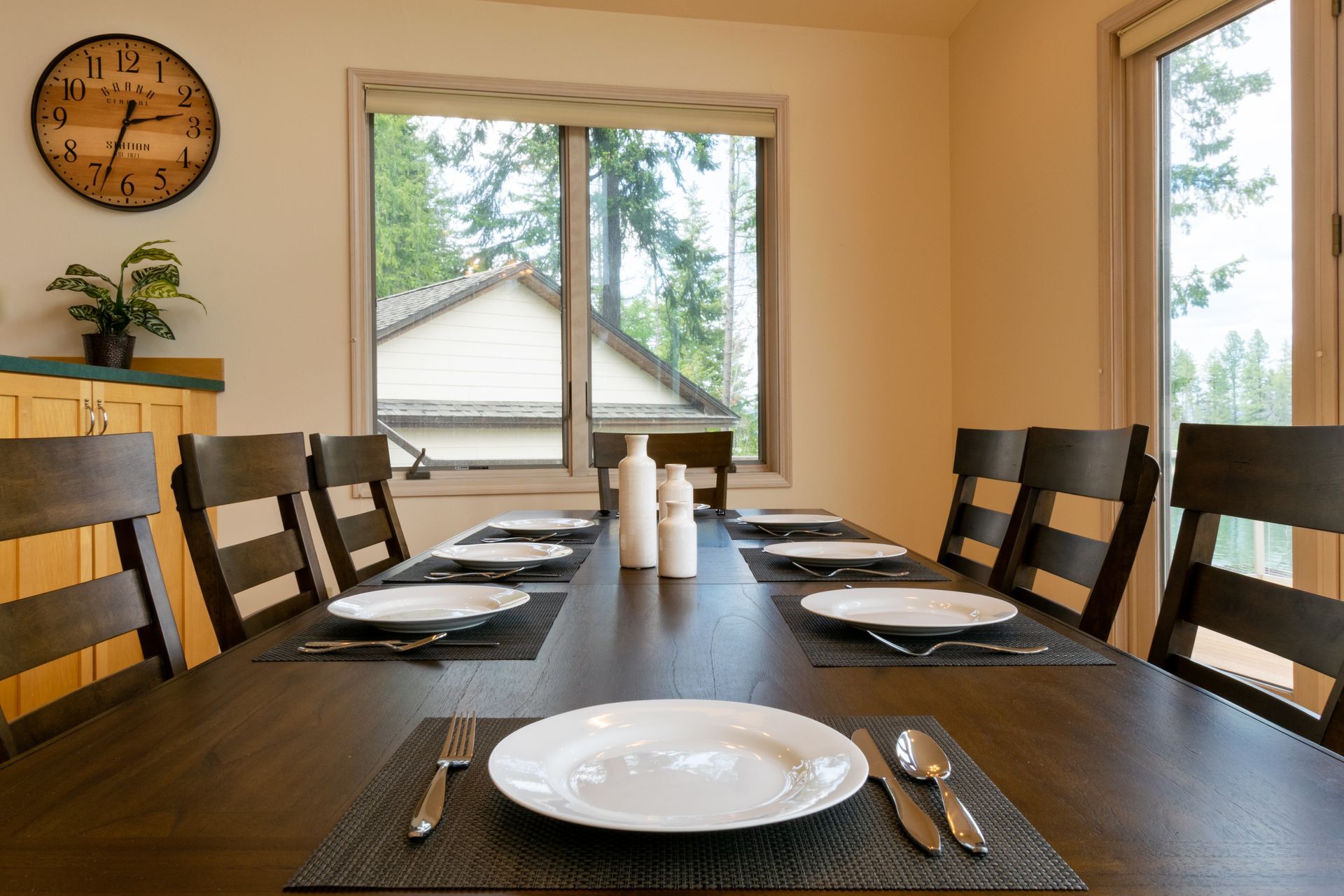 A long dining table set for a meal, with plates, silverware, and centerpieces, by a window overlooking a residential area.