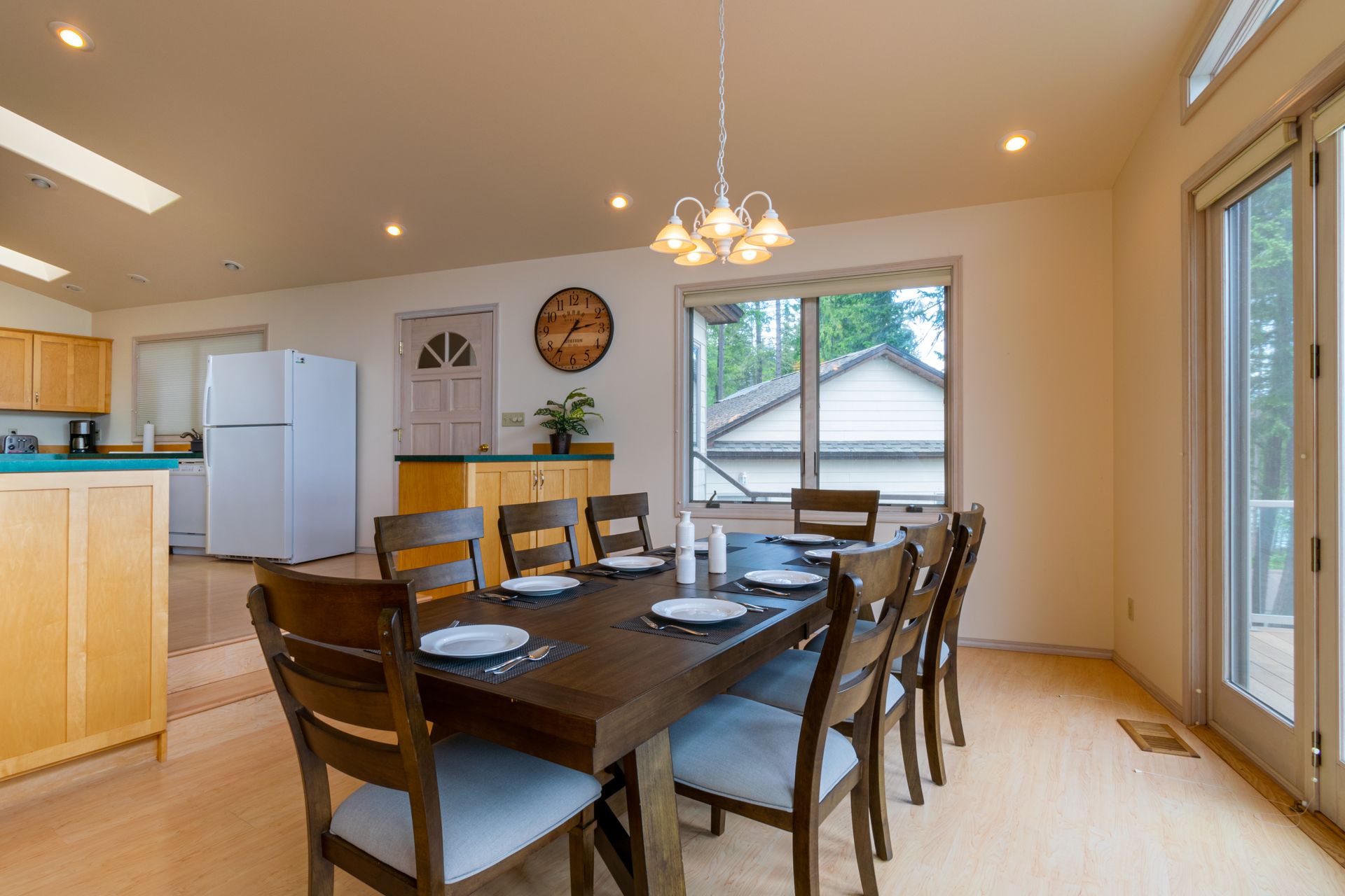 Dining room with large wooden table set for a meal, eight chairs, and a view of outside through a large window.