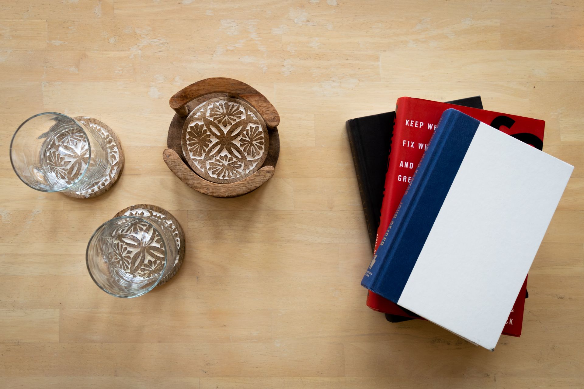 Two glasses and a coaster with decorative designs are next to a stack of books on a wooden surface.