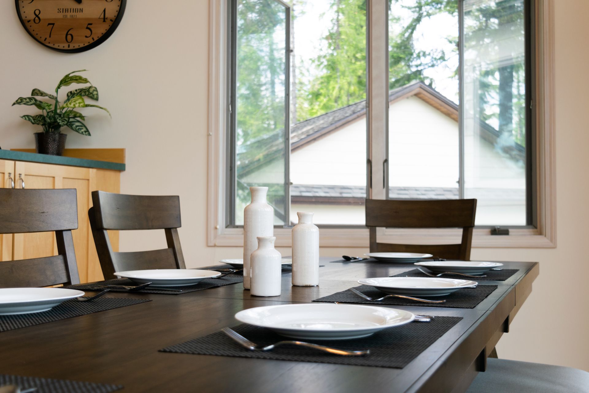 A dining room with a wooden table set for a meal. Sunlight streams through the window overlooking a backyard.