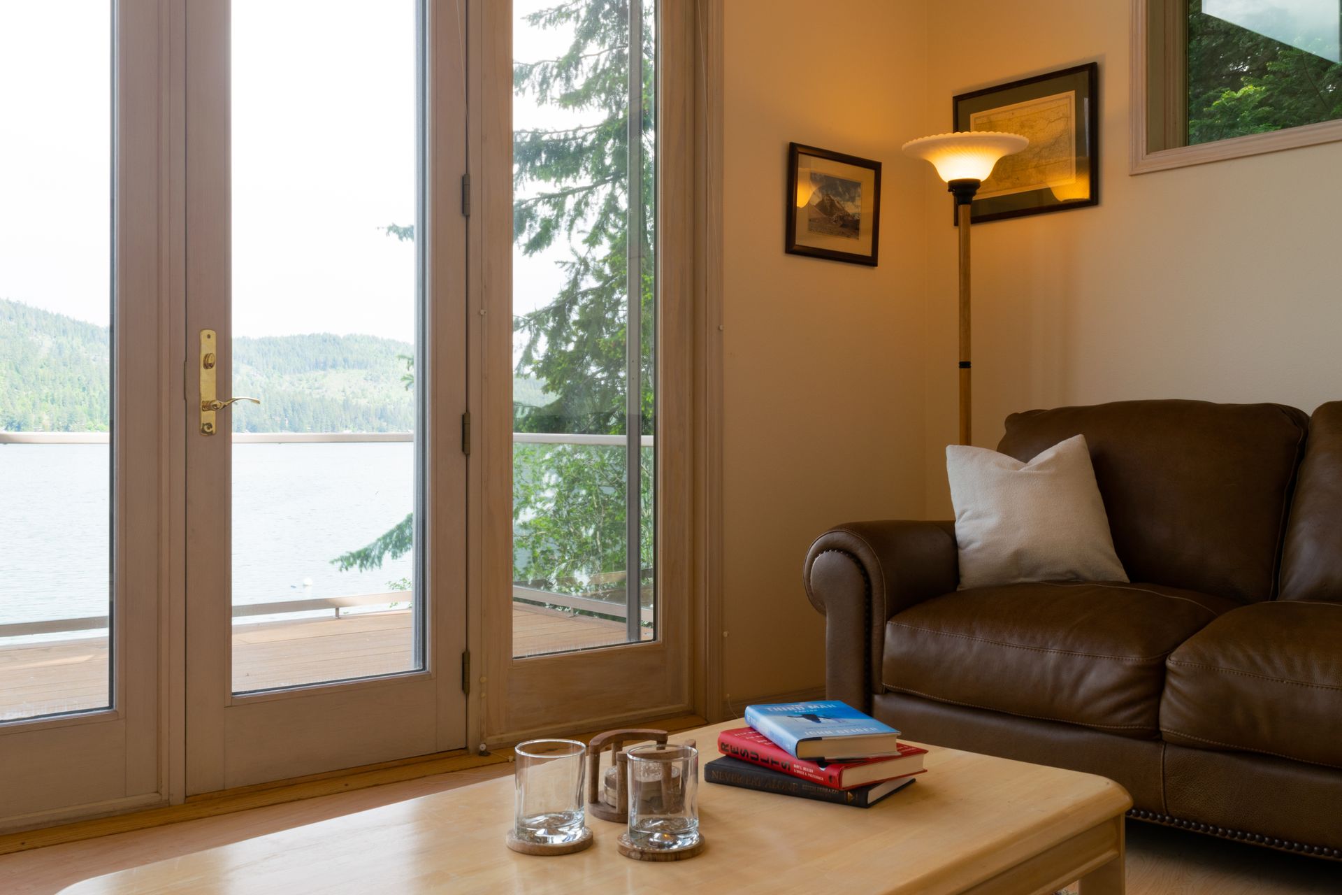 Cozy living room with a leather couch, coffee table with books and glasses, and a view of a lake through glass doors.