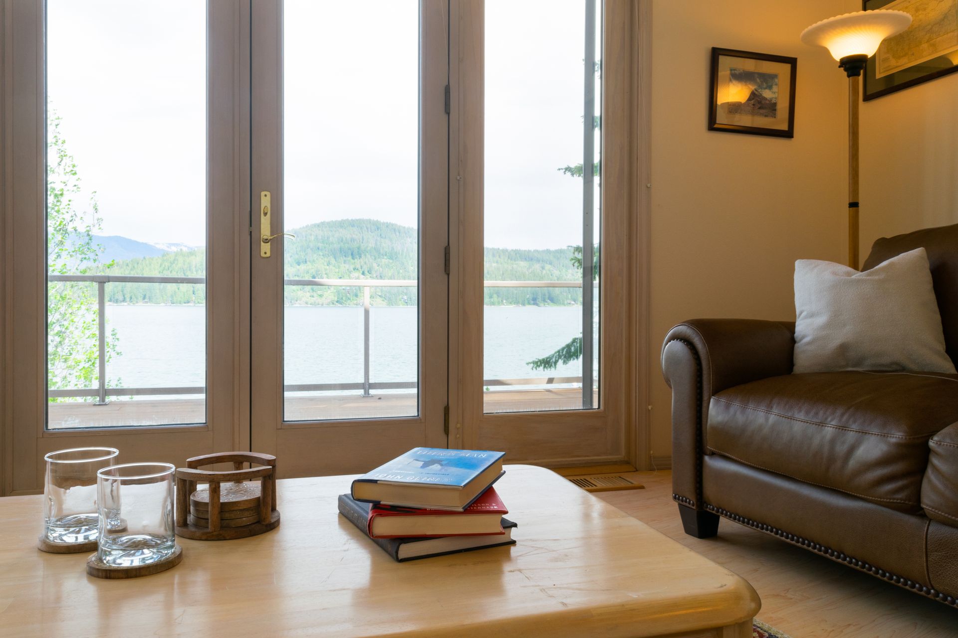 A cozy living room with a lake view. Brown leather sofa, coffee table with books and glasses, and a lamp.