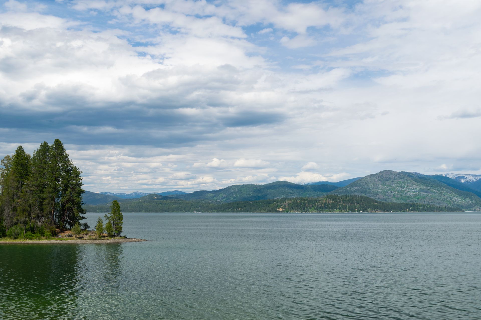 A lake with a forested island in the foreground, mountains in the distance, and a cloudy sky overhead.